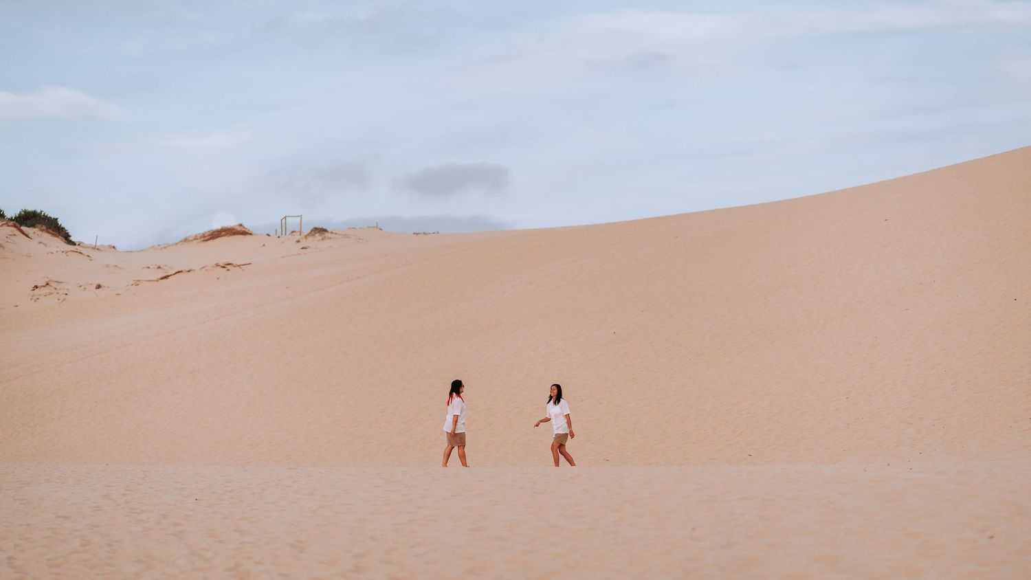 Two distant people walk across a vast sand dune under a pale blue sky in a desert landscape.