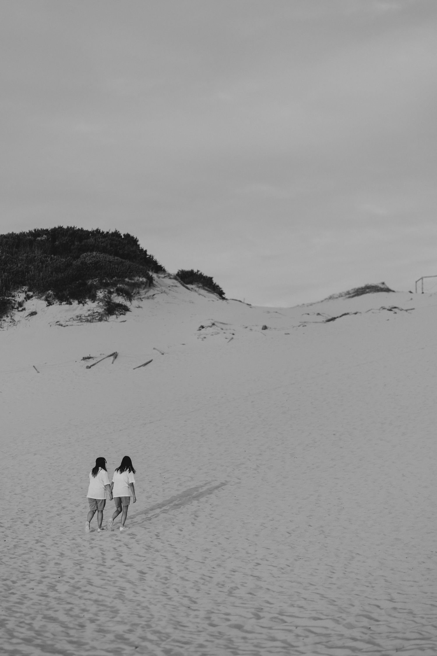 Two figures in white shirts walk together across sweeping sand dunes under an overcast sky in black and white.