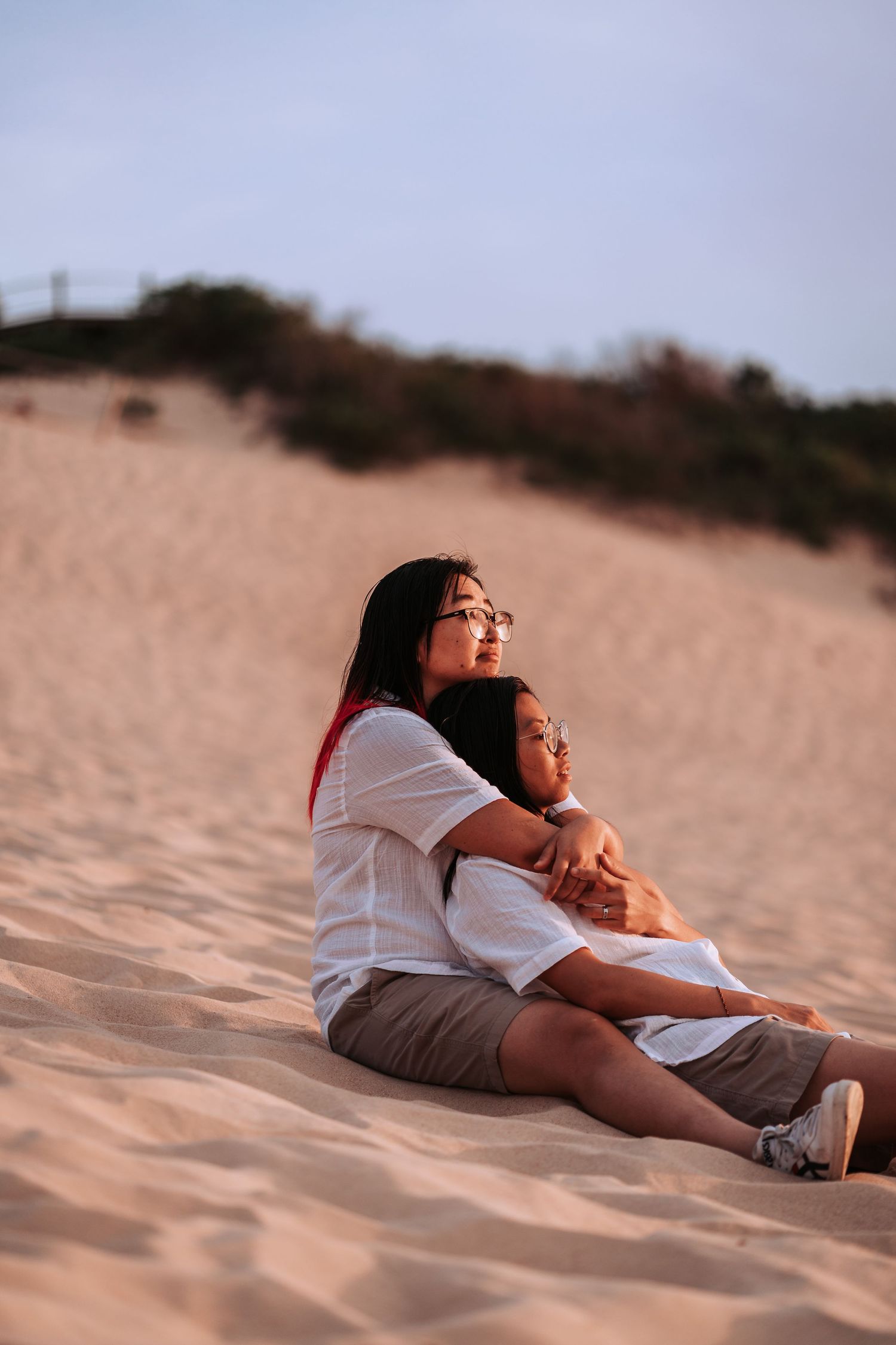 Two individuals wearing white shirts sit together on sandy dunes during golden hour at sunset.