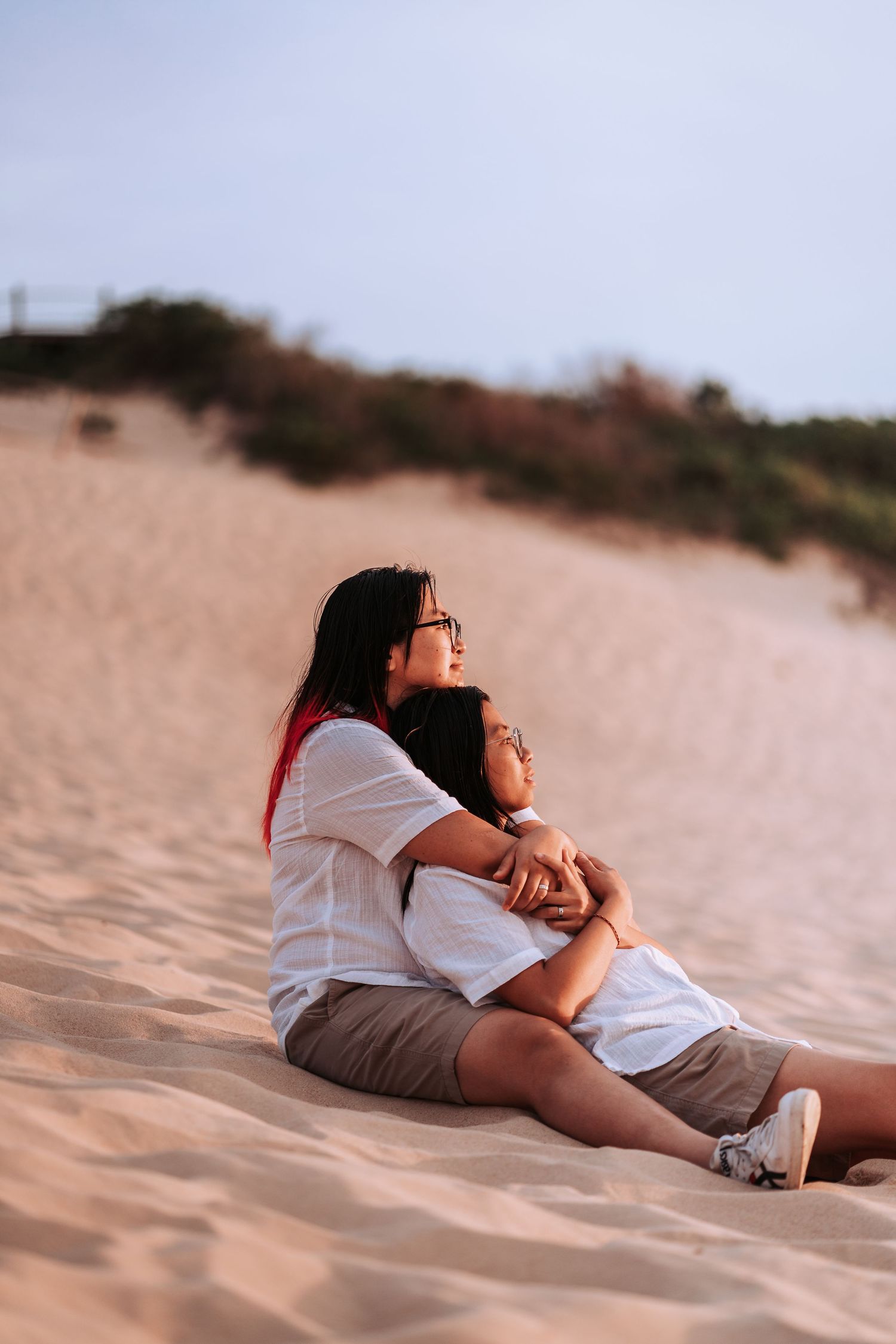 Parent and child in matching white shirts sit together on sandy beach dunes at golden hour.