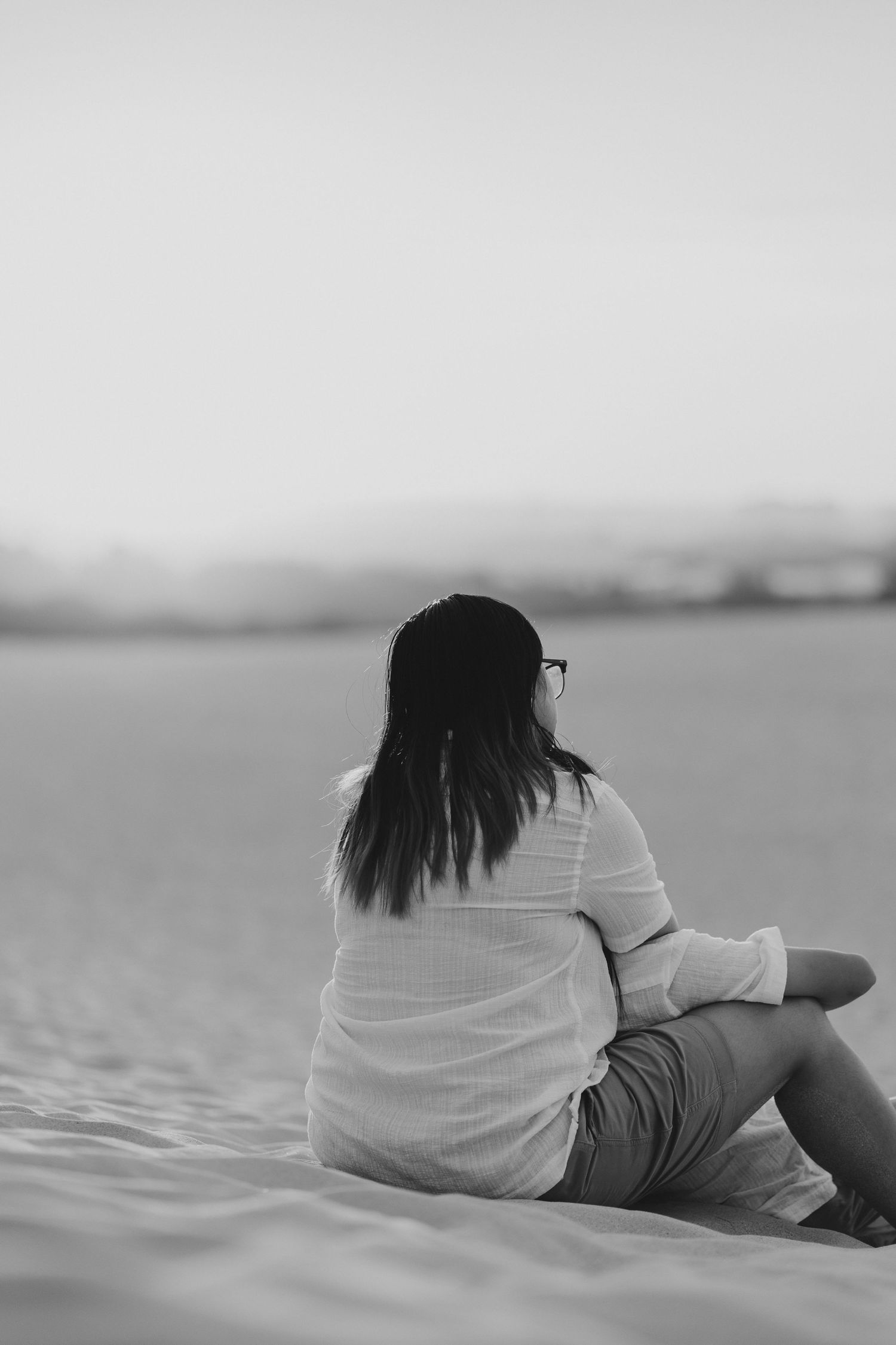 A person with long dark hair sits alone on the beach in a white shirt, facing the ocean in black and white photography.