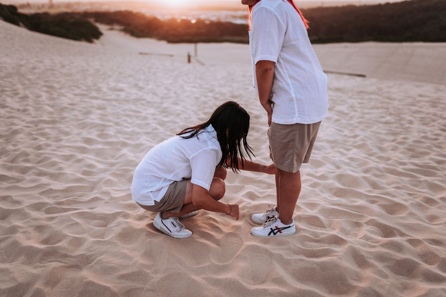 Two people in white clothing on a sandy beach during sunset interacting on the sand.