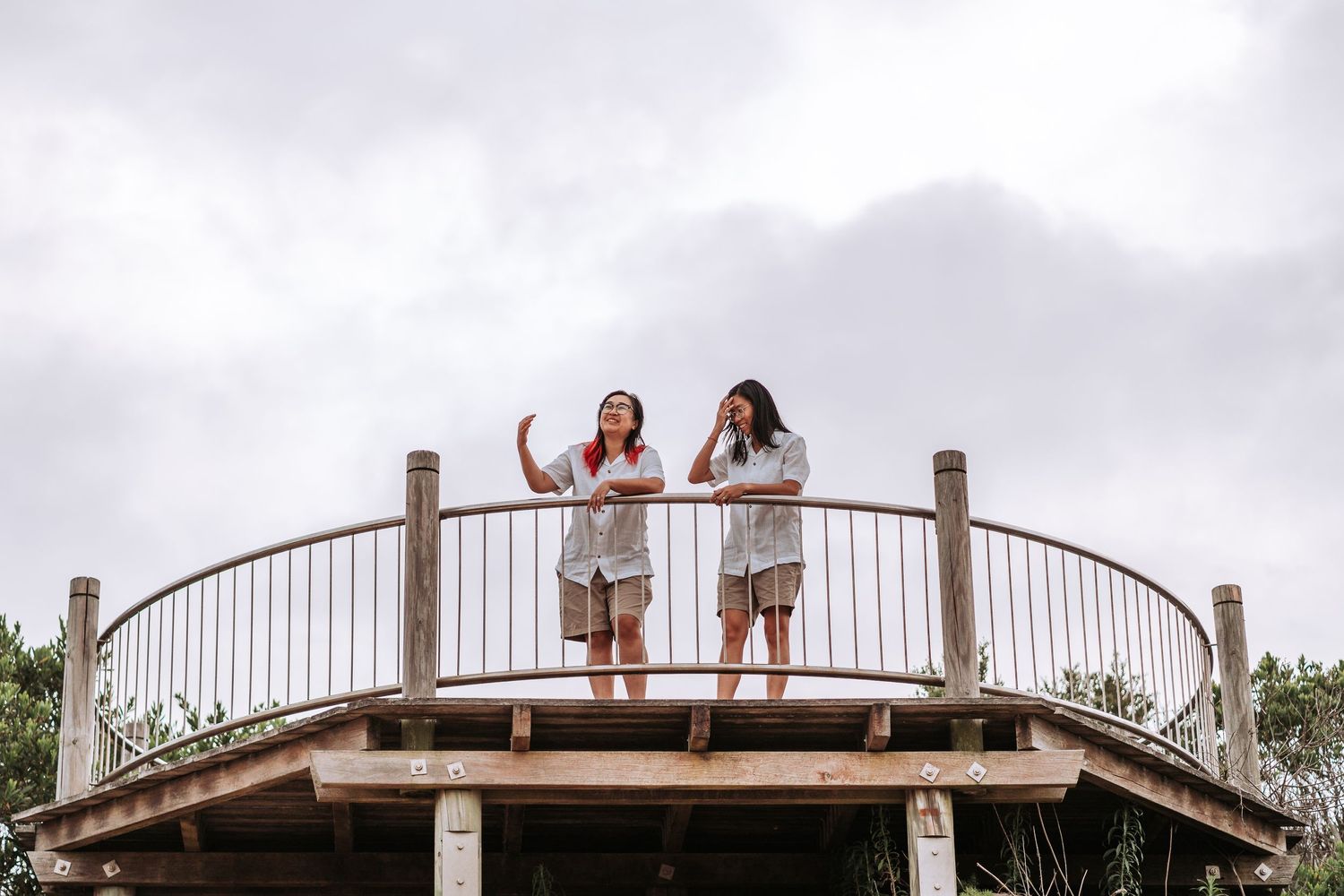 Two people wave while standing on a curved wooden observation deck on an overcast day.