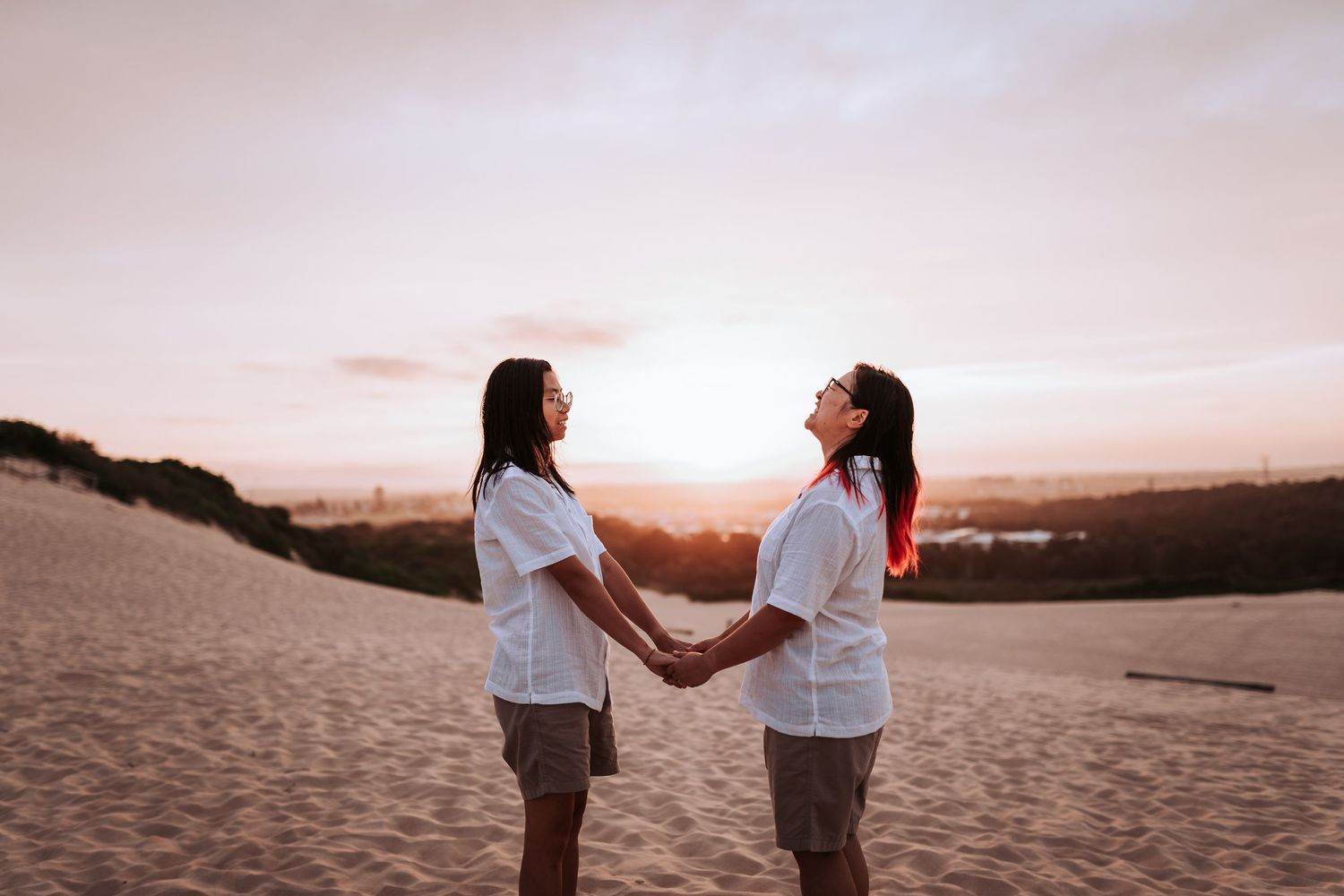Two people in white shirts hold hands on sandy dunes during a warm sunset.