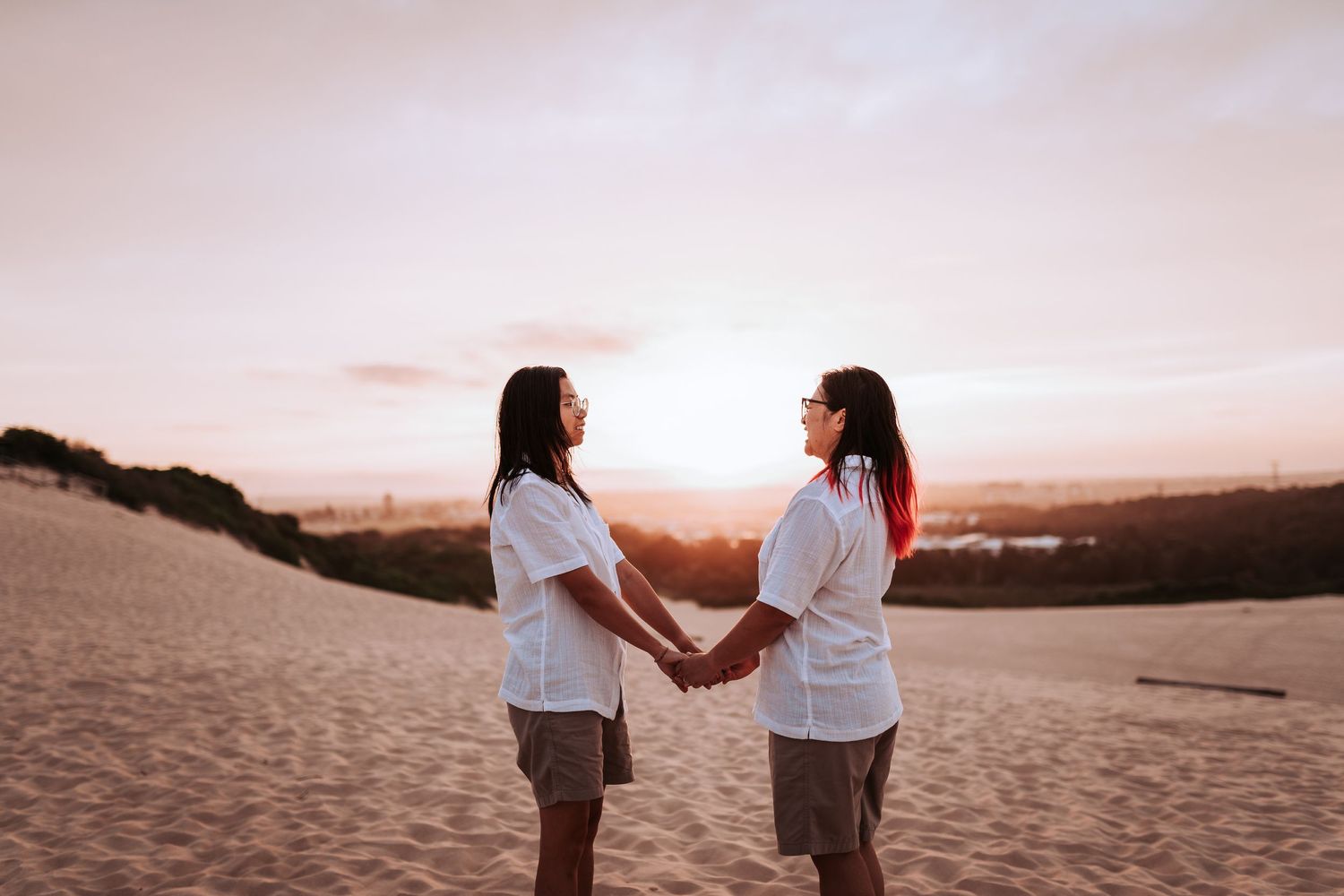 Two people in white shirts hold hands at sunset on a sandy hill overlooking a distant landscape.