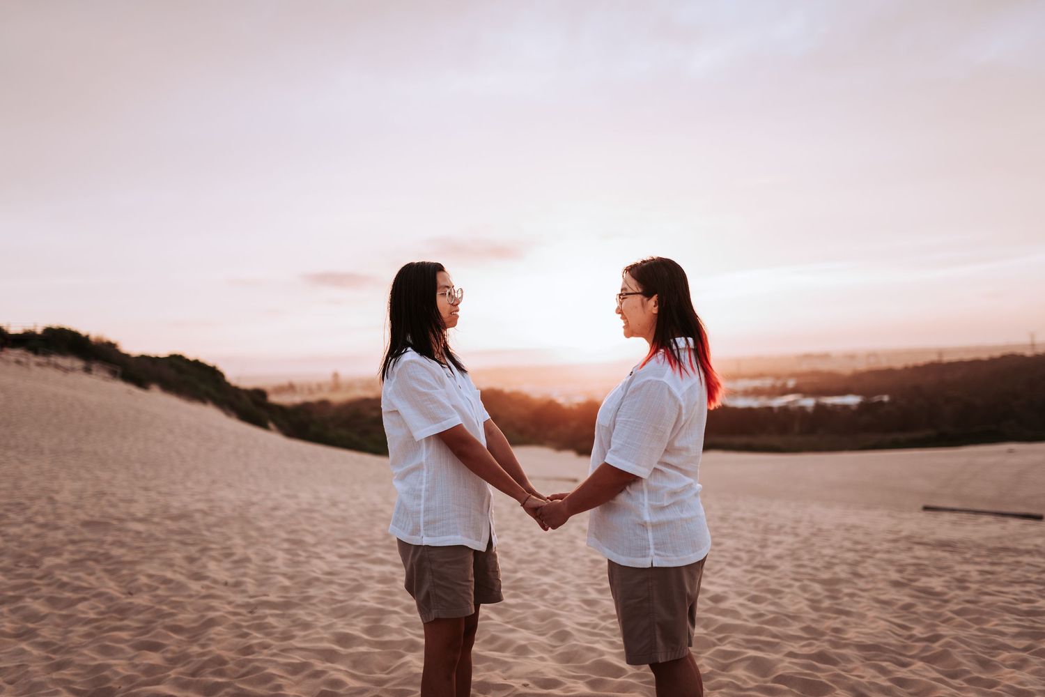 Two people in white shirts hold hands while standing on sandy dunes at sunset.