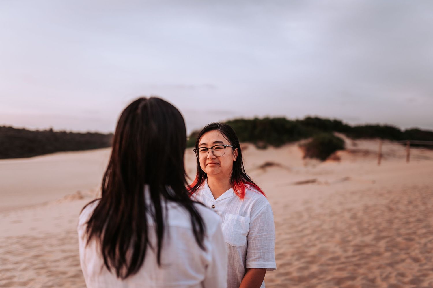 Two people in white shirts share an intimate moment while standing on sandy dunes at dusk.