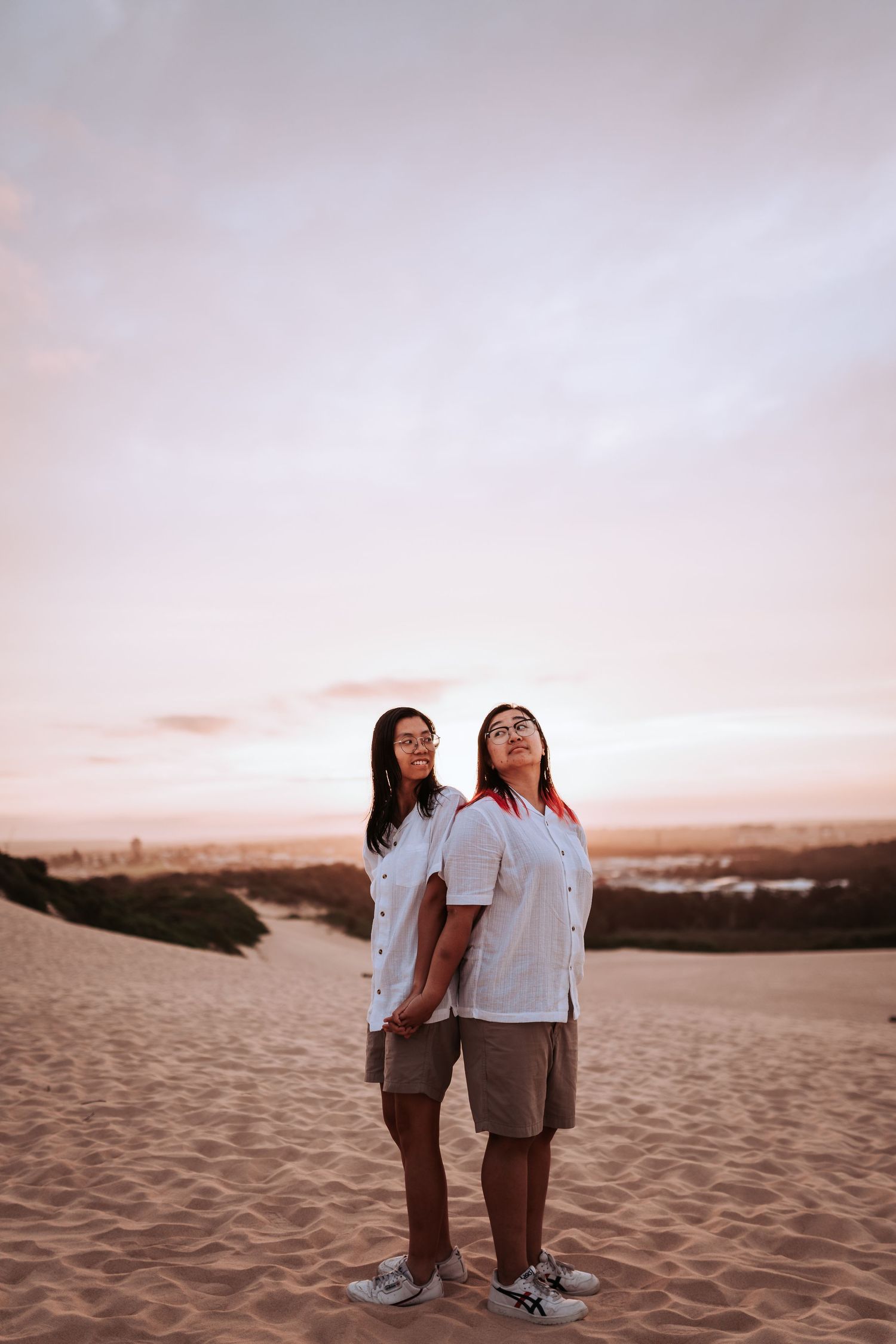 Two people in matching white shirts stand together on sandy dunes at sunset with a soft pink sky in the background.