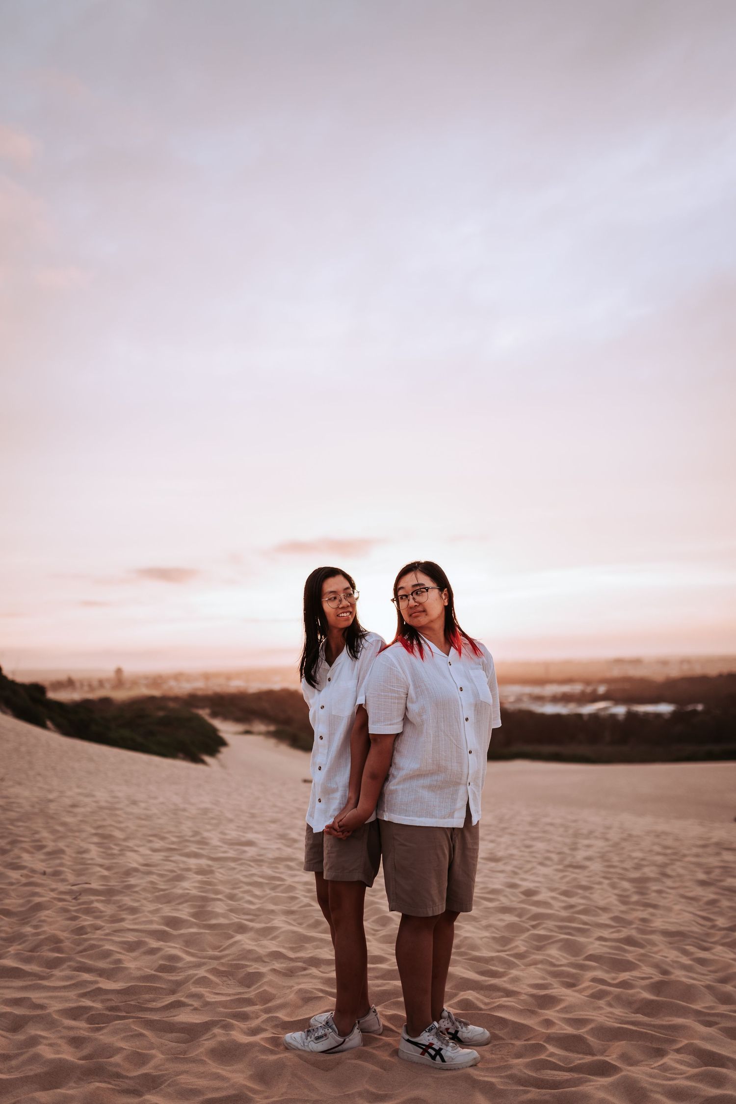 Two people wearing matching white shirts and khaki shorts stand together on sand dunes at sunset.