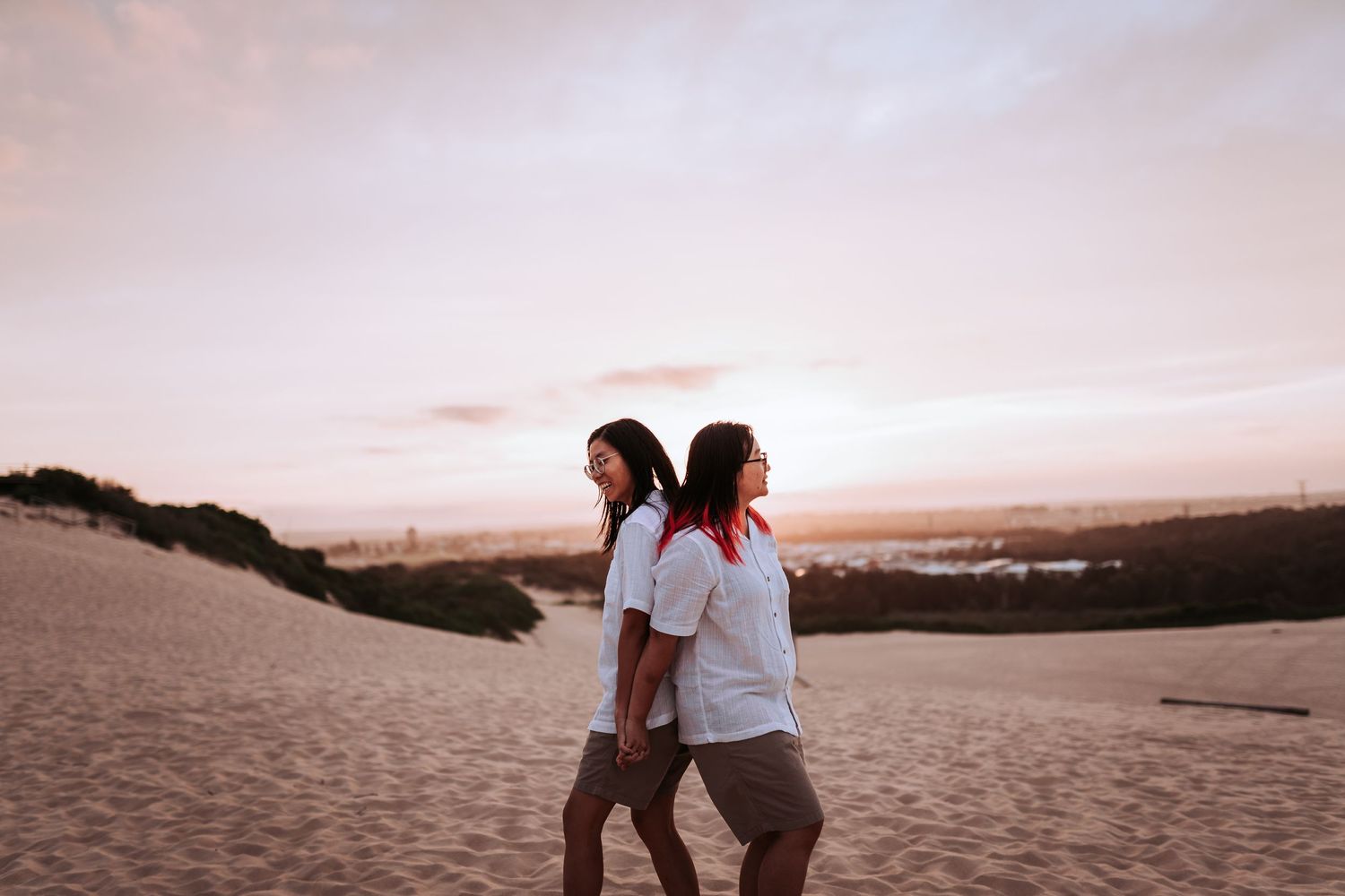 Two people walk together on sand dunes during a colorful sunset with hills visible in the distance.