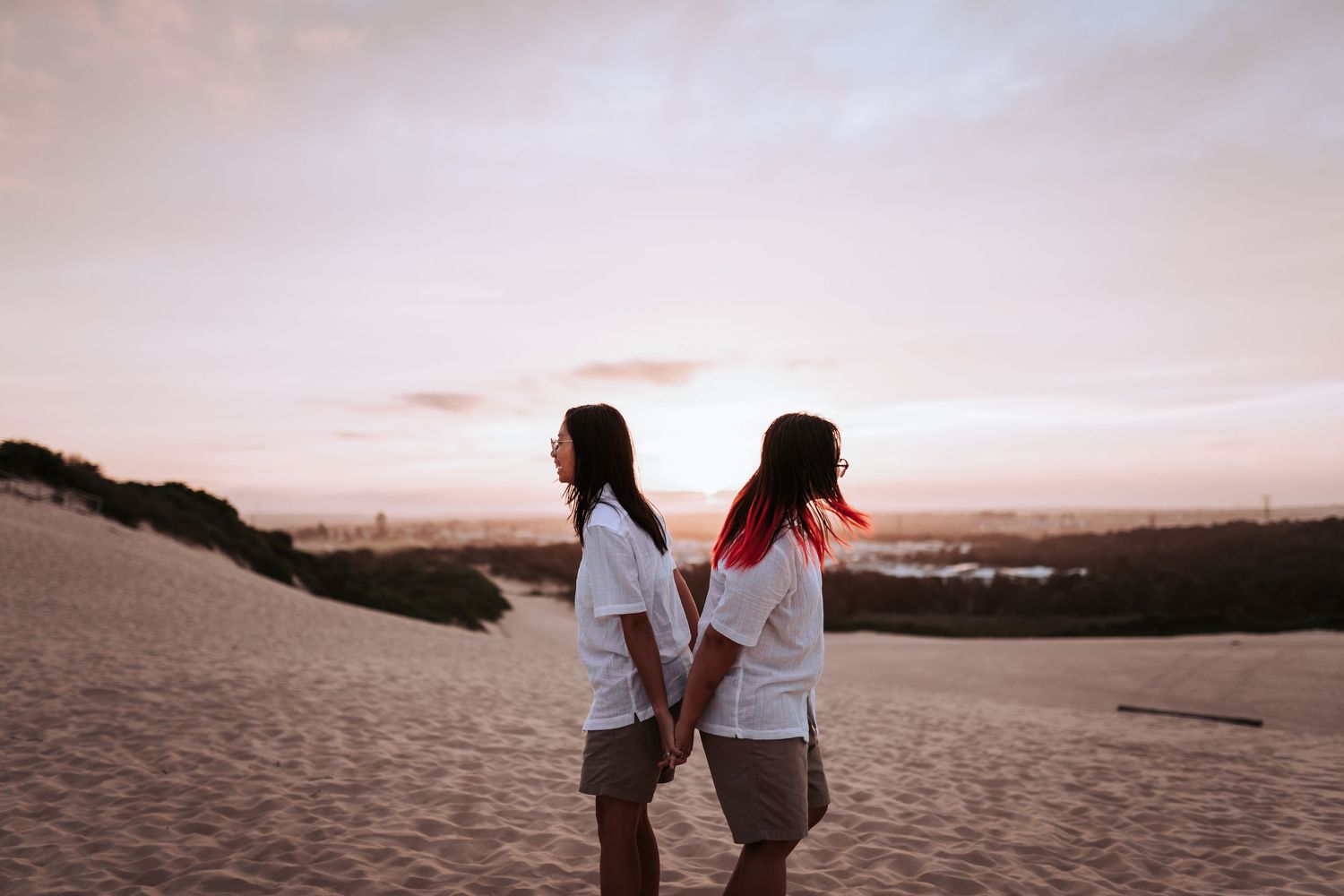Two people in white shirts standing back to back on sandy dunes at sunset with pink sky in background.