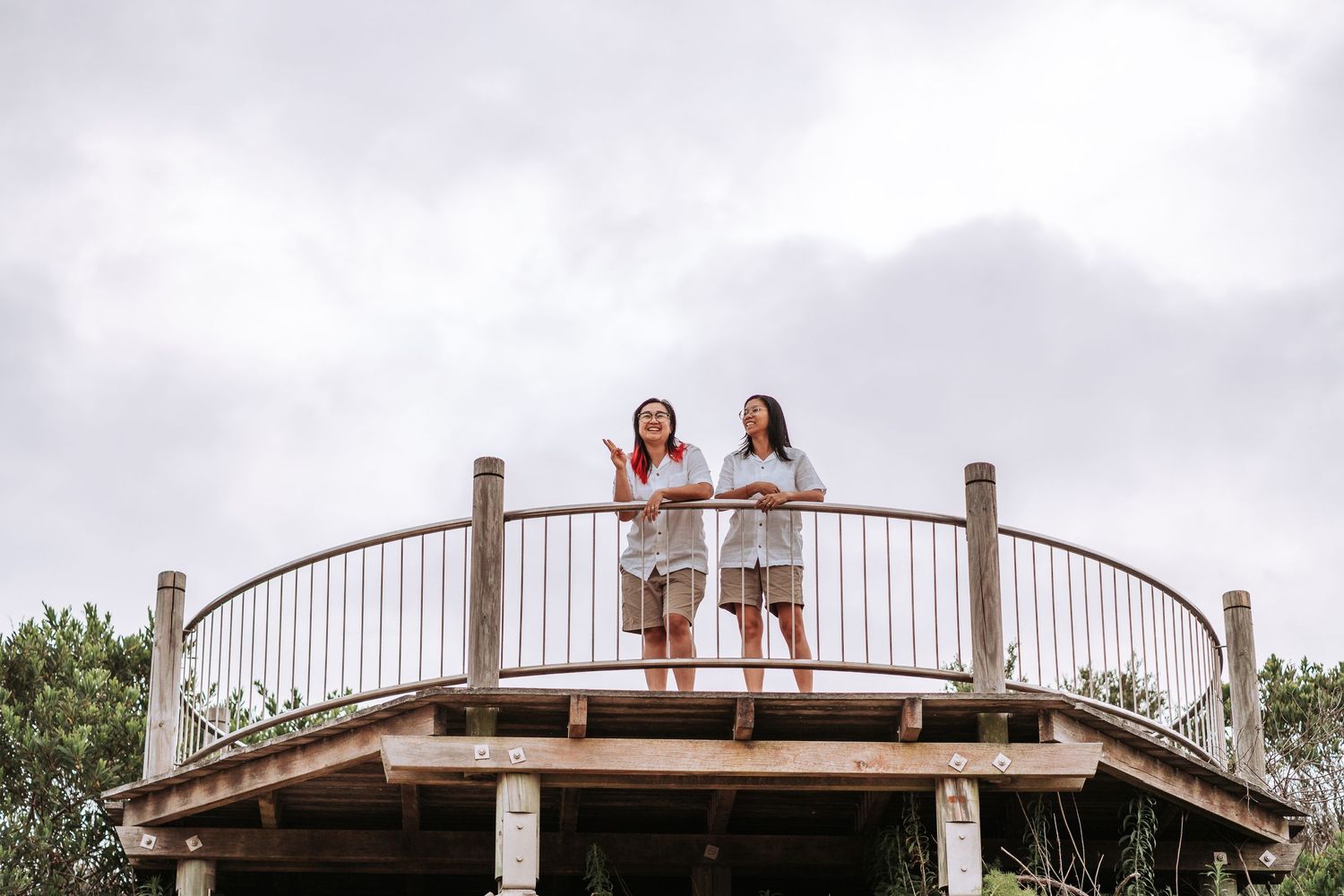 Two women in white outfits stand on a curved wooden observation deck against a cloudy sky and green trees.