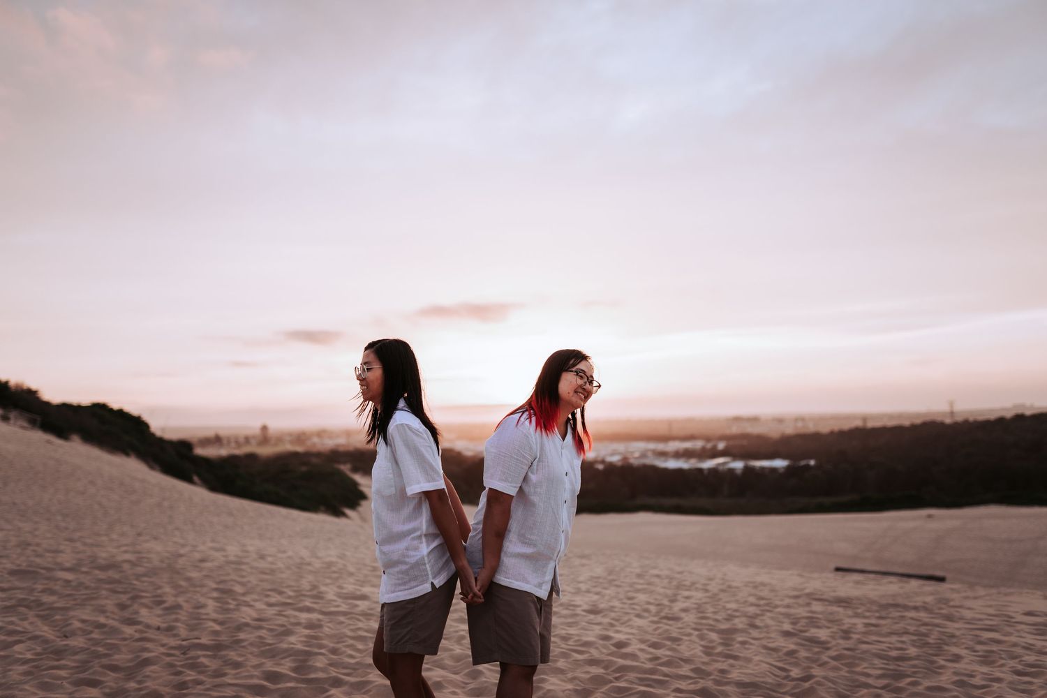 Two people in white shirts walking together on sandy dunes during sunset with distant city views in background.