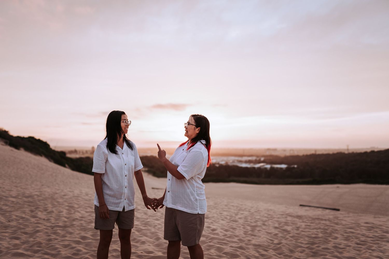 Two people share a joyful moment while standing on desert dunes at dusk with a pink sky backdrop.