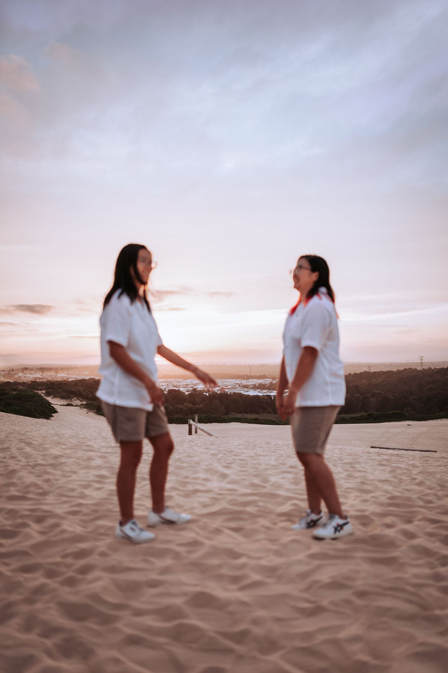 Two people in white shirts and beige shorts stand together on a sandy beach during sunset.
