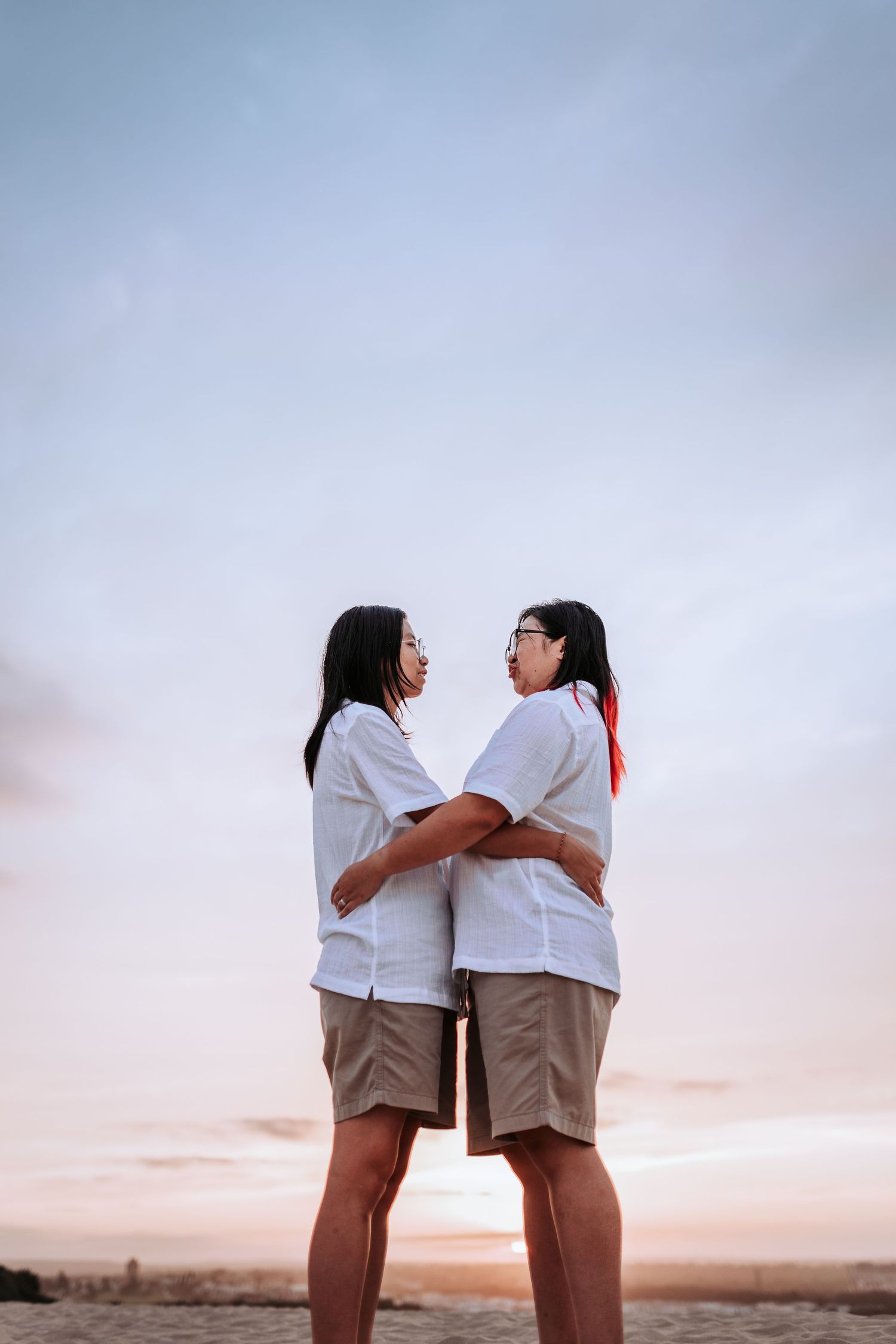 Two people in matching white shirts and khaki shorts embrace while watching the sunset together against a soft sky.