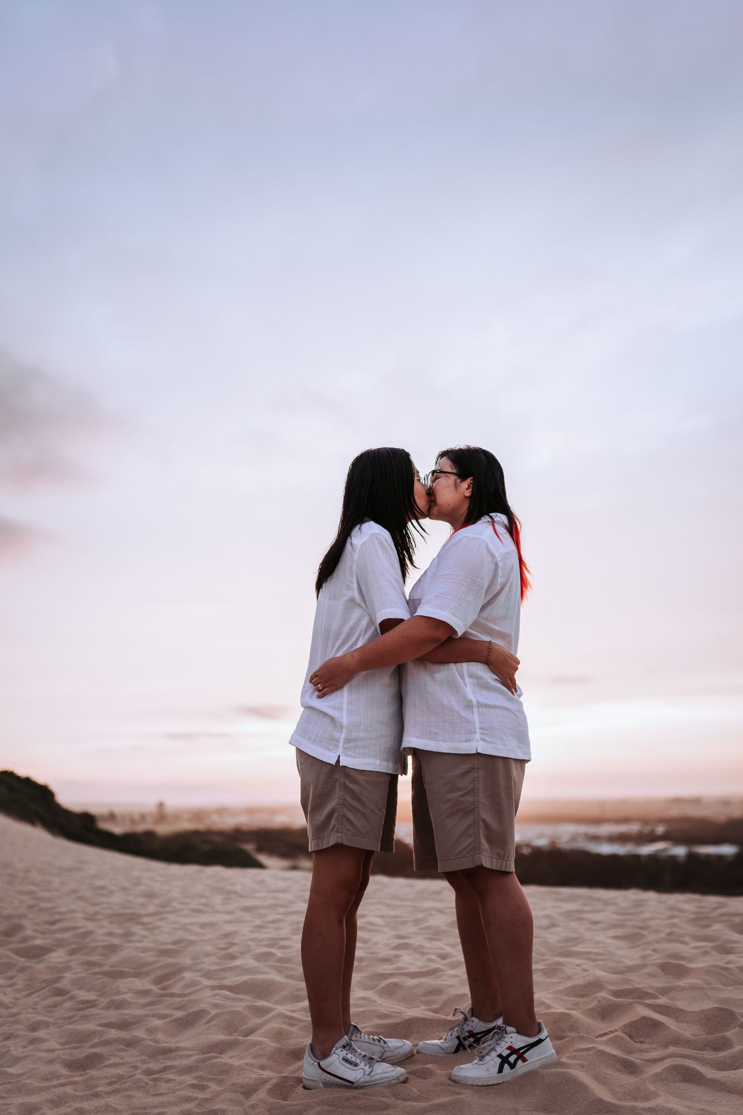 A romantic couple shares a tender kiss on a sandy beach at sunset while wearing matching white shirts and shorts.