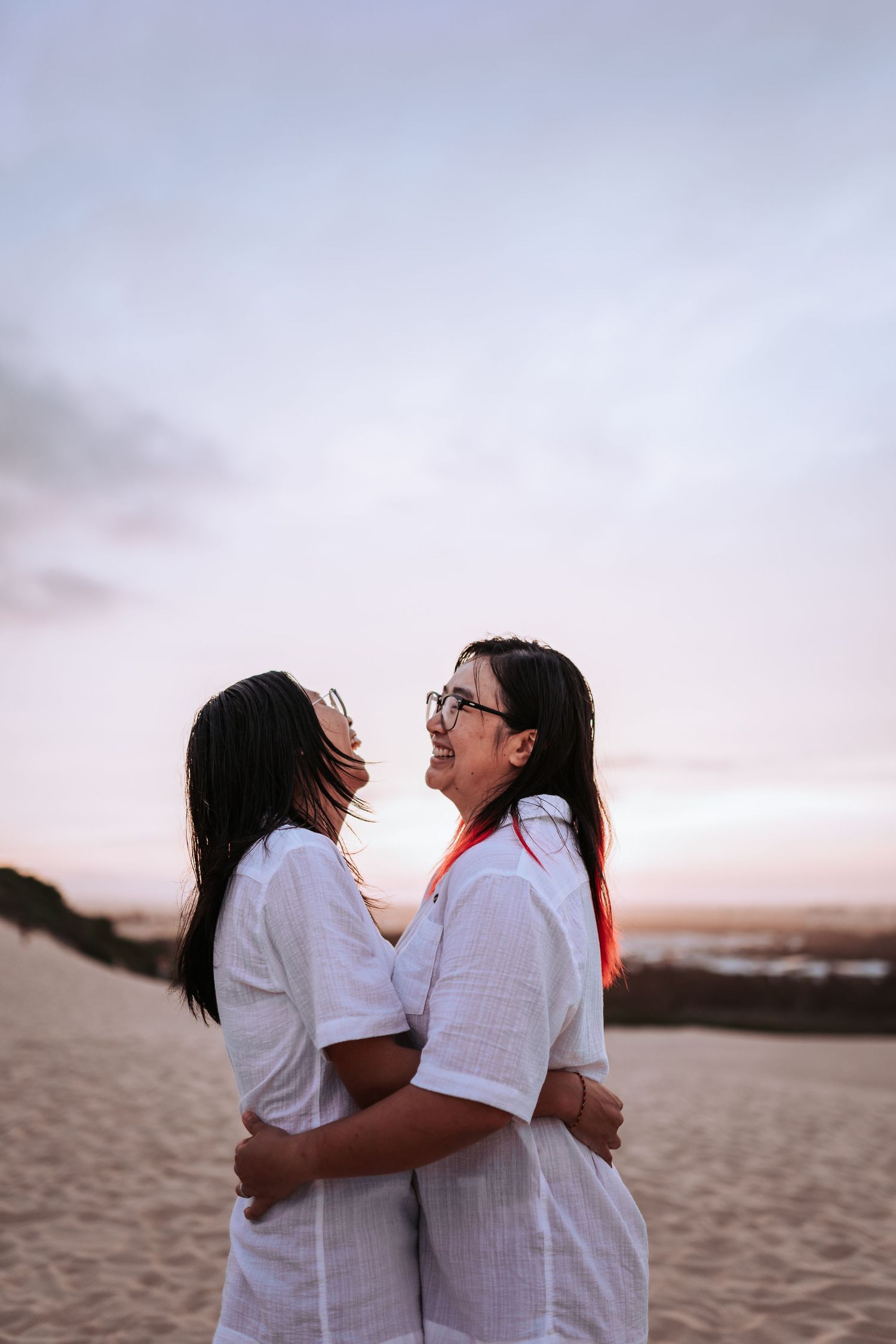 Two people wearing white shirts laugh together while embracing on a beach at sunset.