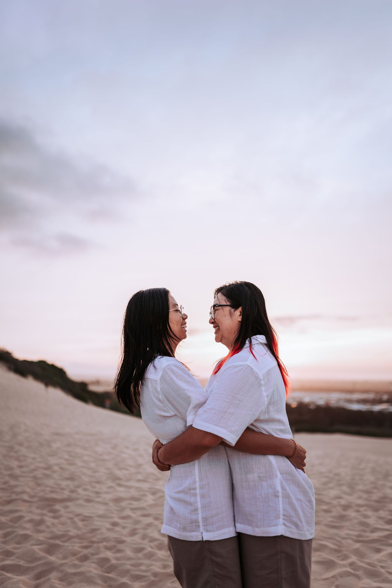 A couple in matching white shirts embrace and laugh together on a sandy beach at sunset.