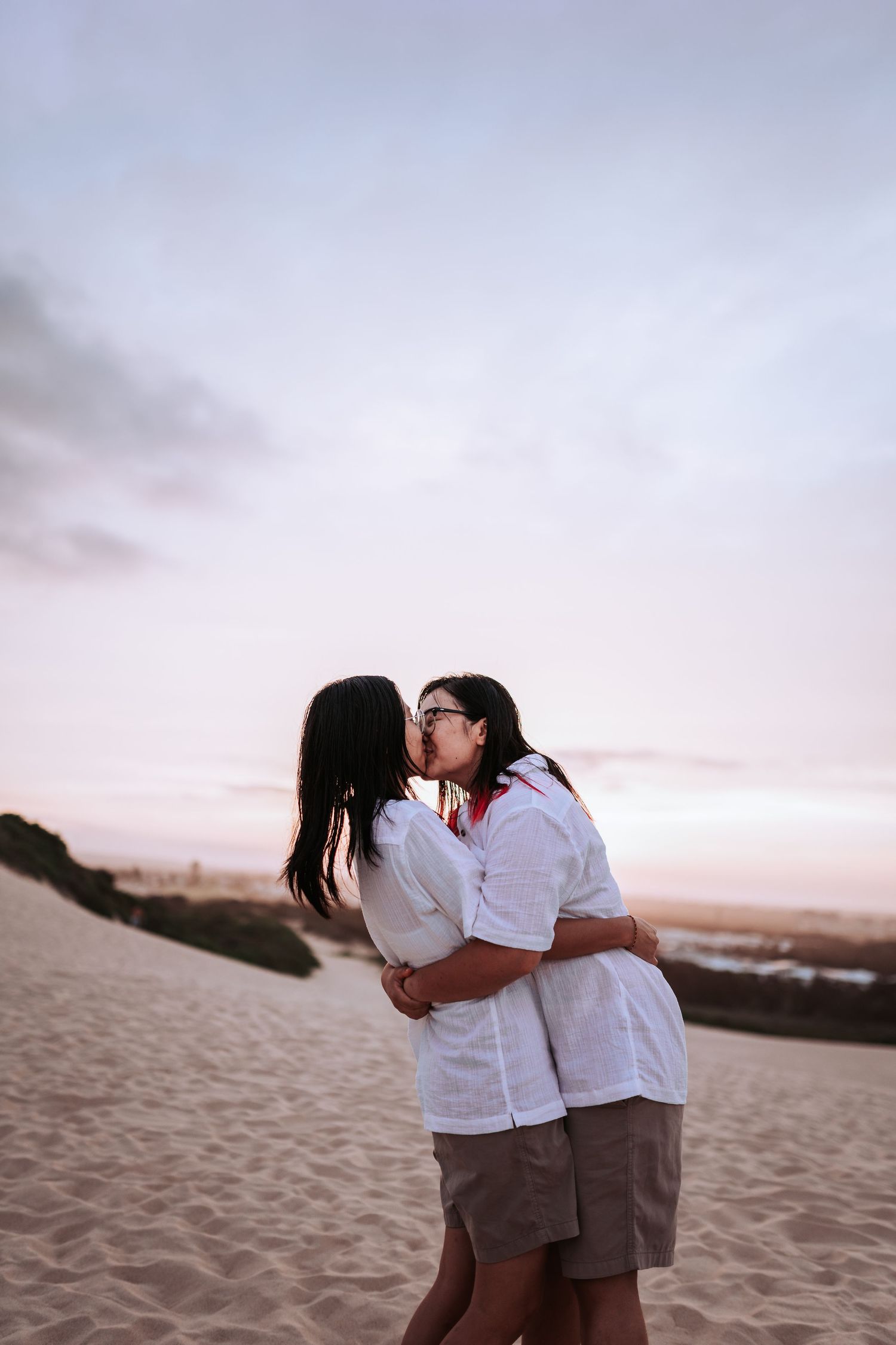 A couple in matching white shirts share an intimate moment on a sandy beach at sunset.