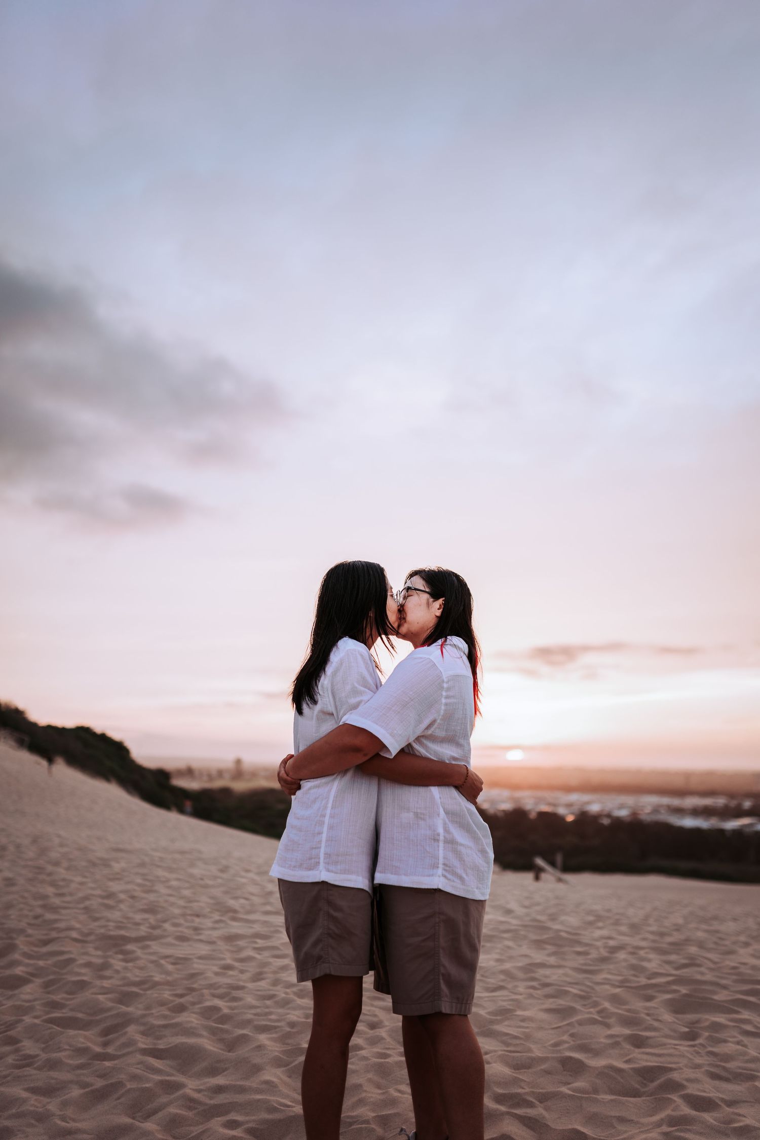A couple in matching white shirts and khaki shorts embraces on a sandy beach at sunset.