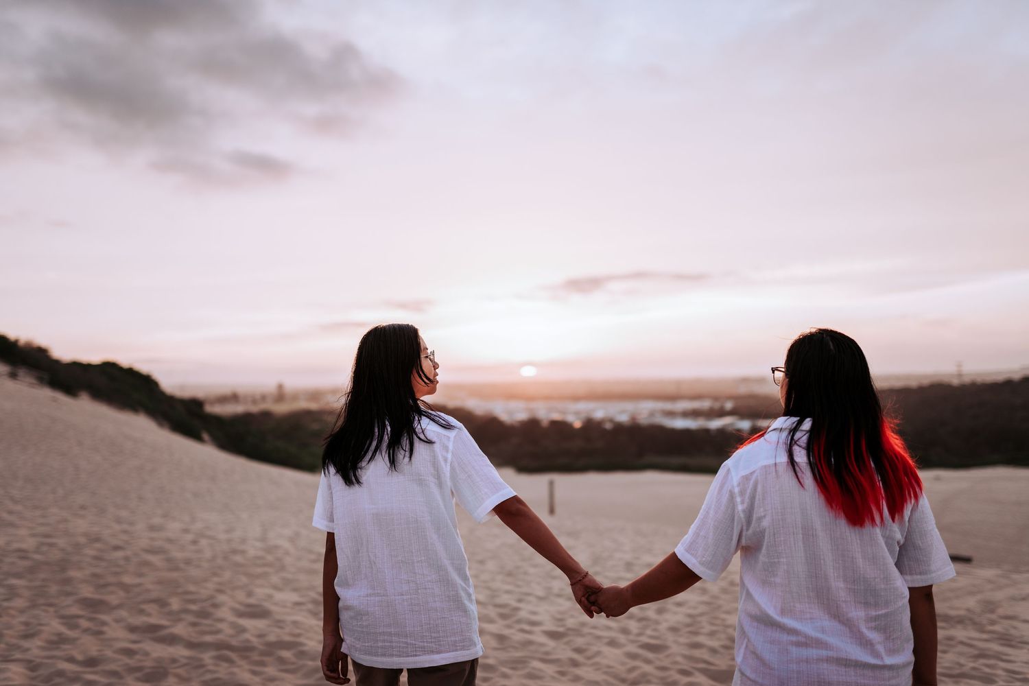 Two people wearing white shirts hold hands while watching sunset together on sandy beach.