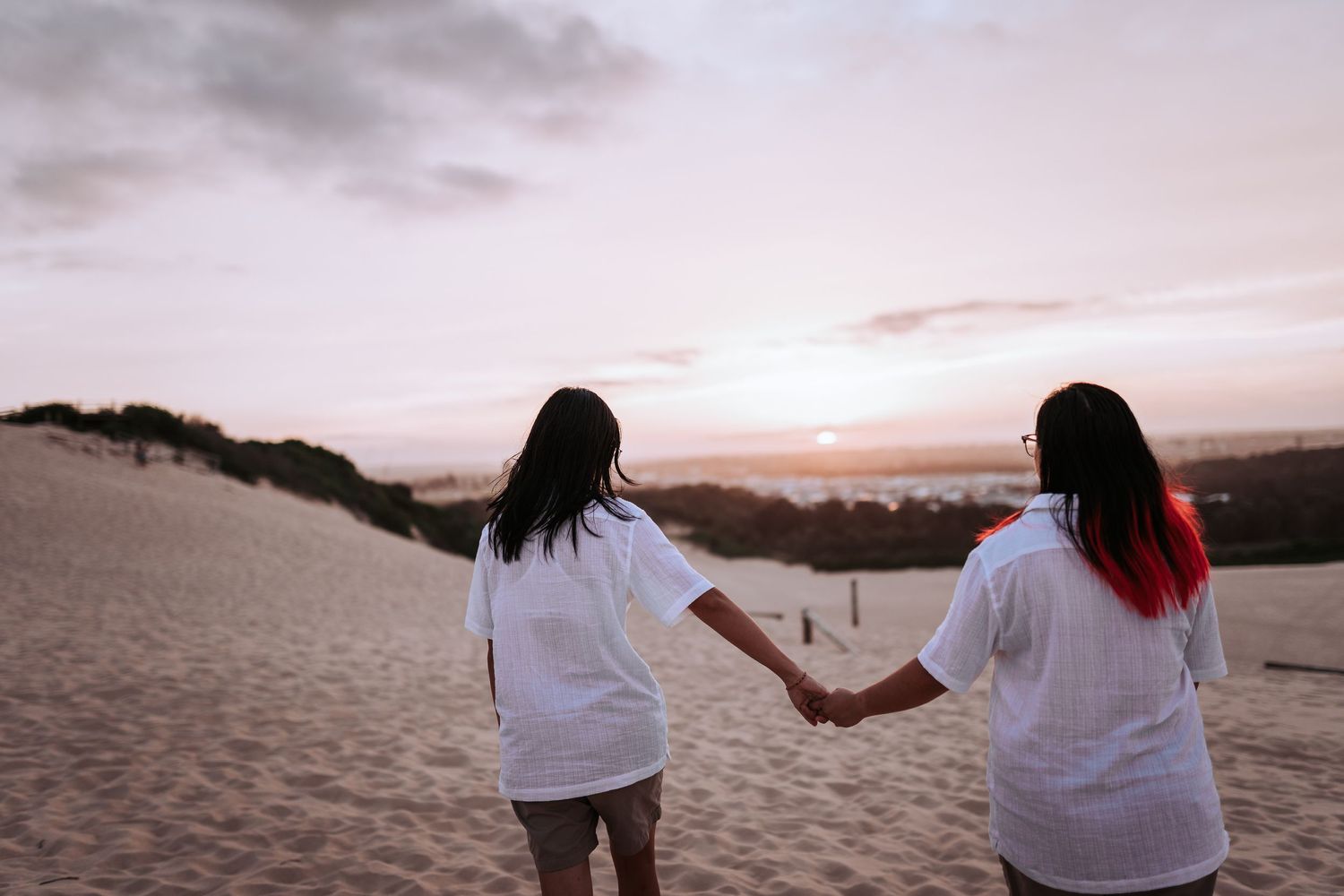 Two people in white shirts holding hands while walking on sand dunes at sunset.