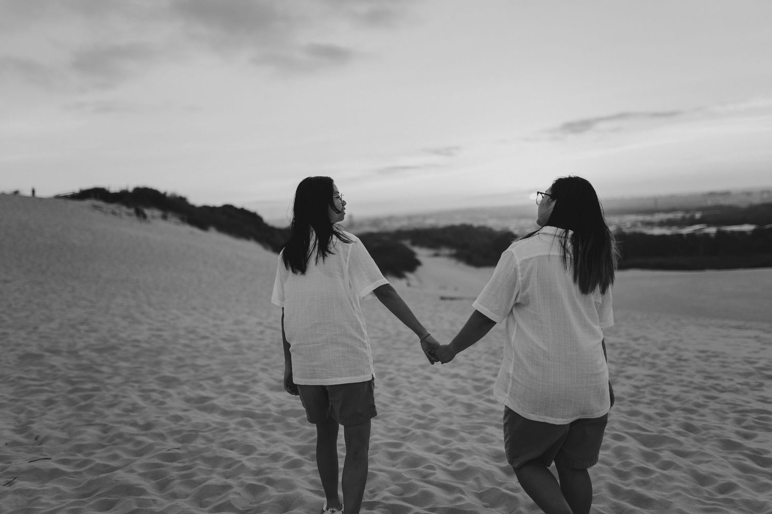 Two people in white shirts holding hands while walking together on sand dunes at sunset in black and white.