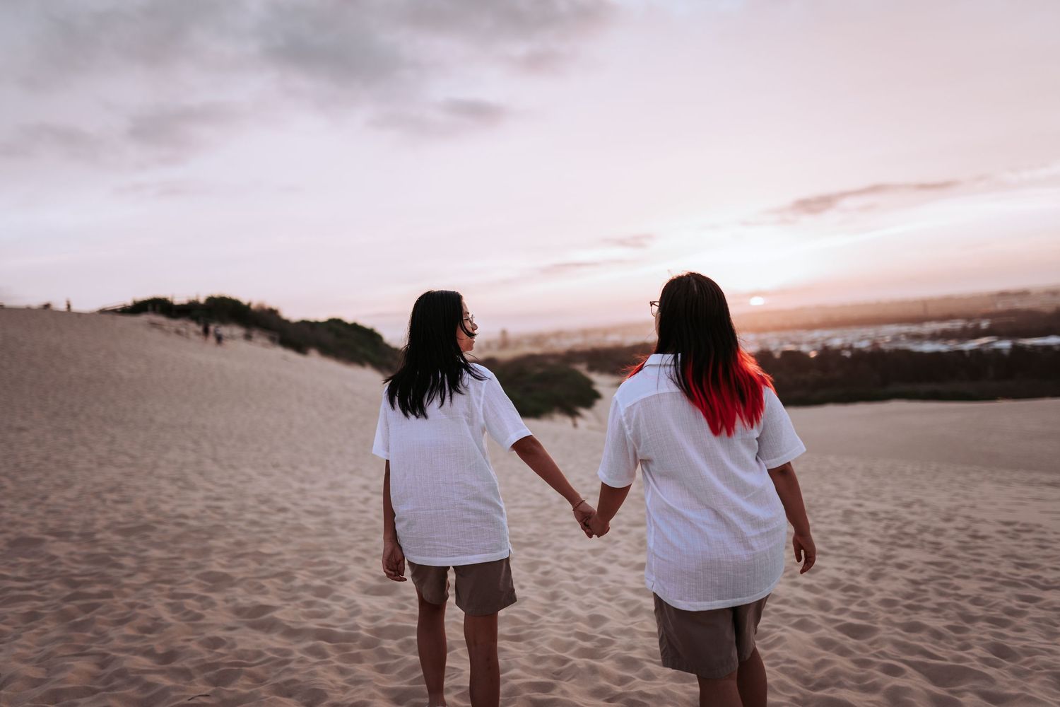 Two friends holding hands walk on sandy beach at sunset wearing white shirts against pink and purple sky.