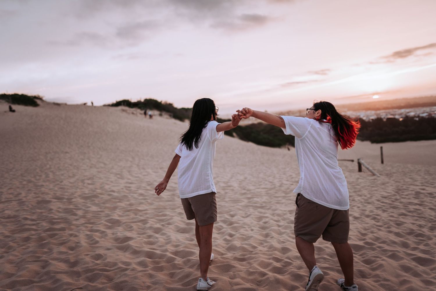 Two people in white t-shirts hold hands while walking through sand dunes at sunset.