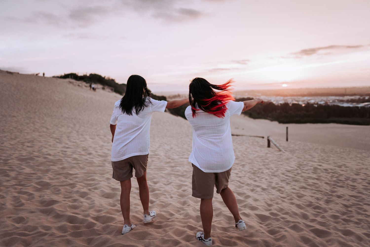 Two people in white shirts run together on sand dunes at sunset.