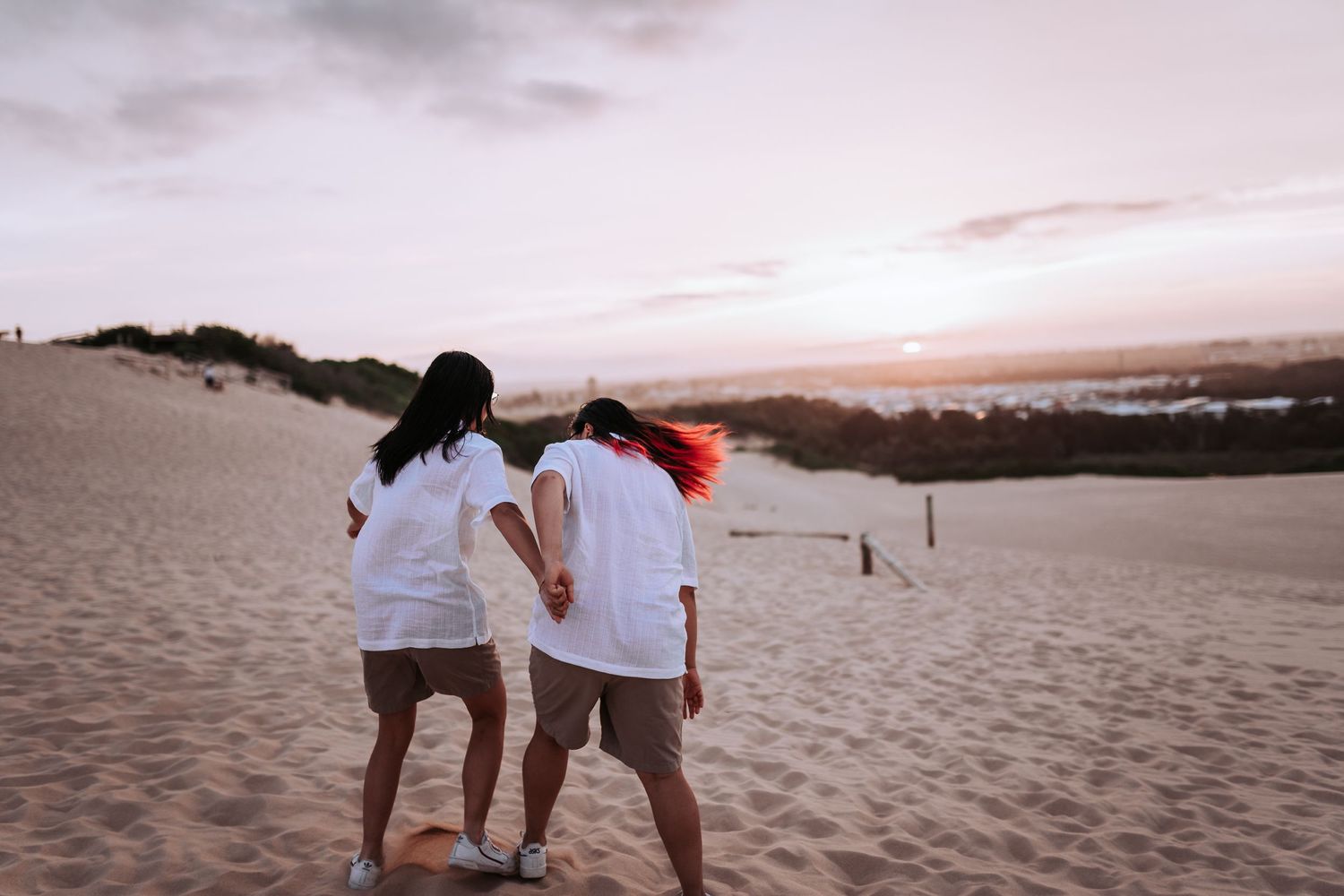 Two people stand together on sandy dunes overlooking the ocean at sunset, creating a romantic silhouette.