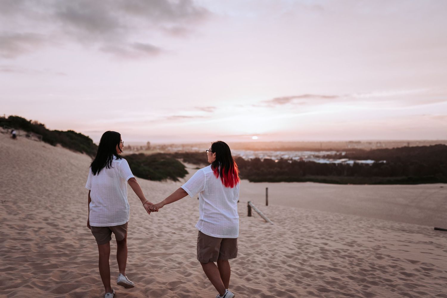 Two people in white shirts holding hands while walking on sand dunes at sunset.