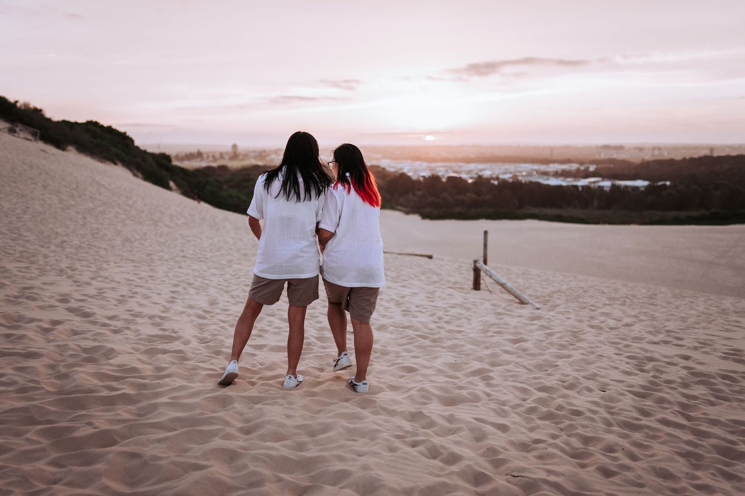 Two people in white shirts stand together on sand dunes watching a pink sunset over the ocean.