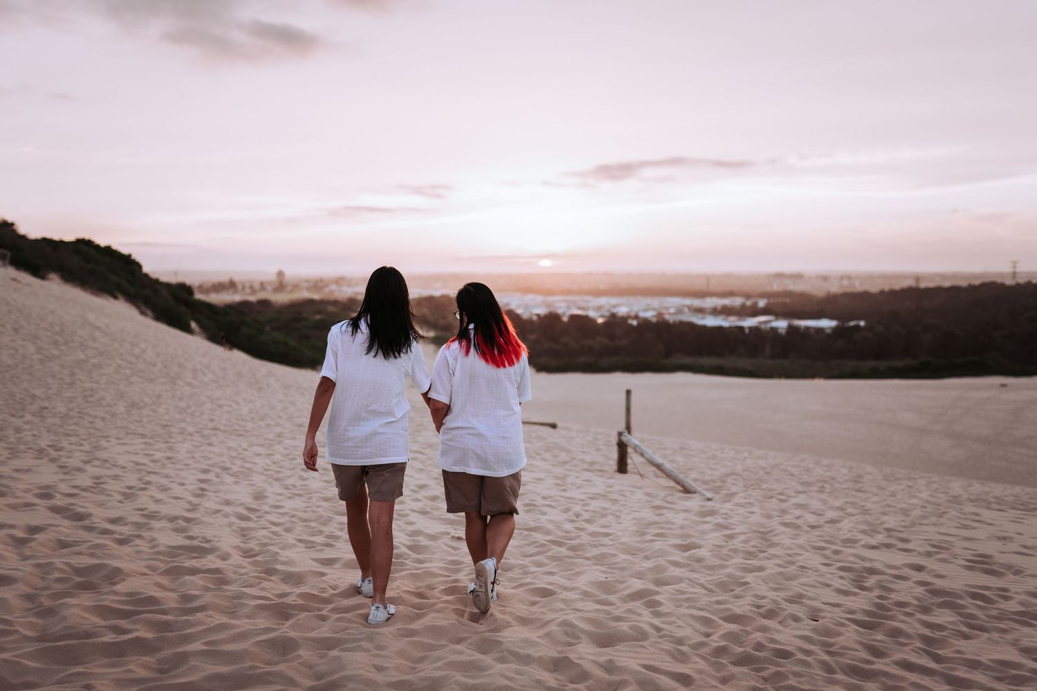 Two people walking together on sandy dunes at sunset overlooking a coastal view.