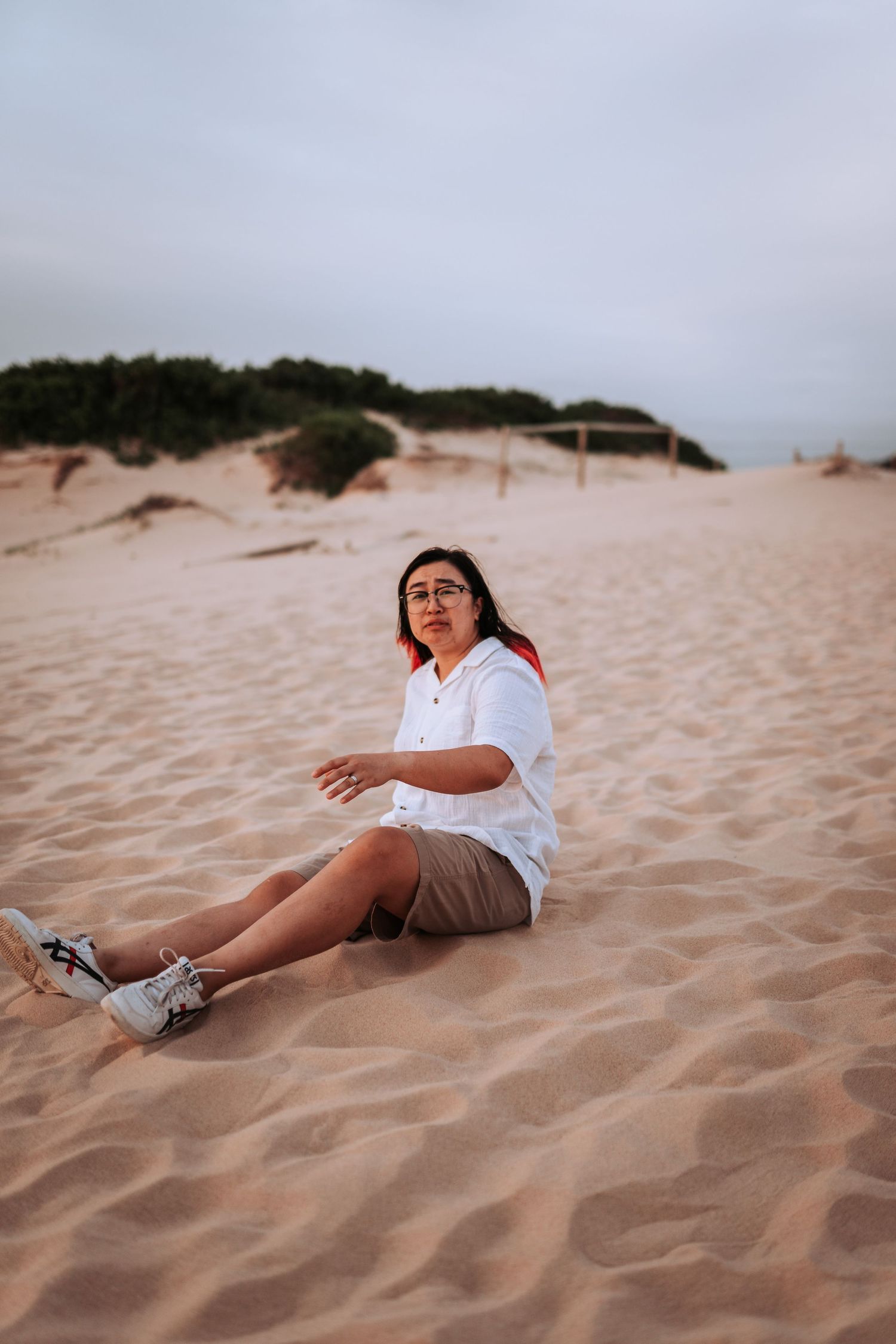 Someone poses casually sitting on sandy dunes at sunset wearing a white shirt and brown shorts.
