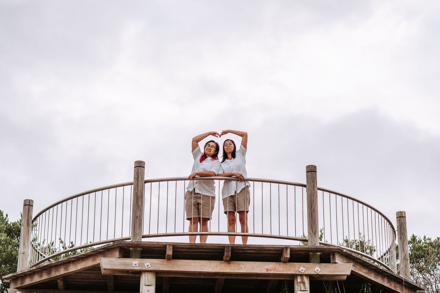 Two figures in matching outfits stand together on a curved wooden observation deck against a cloudy sky.