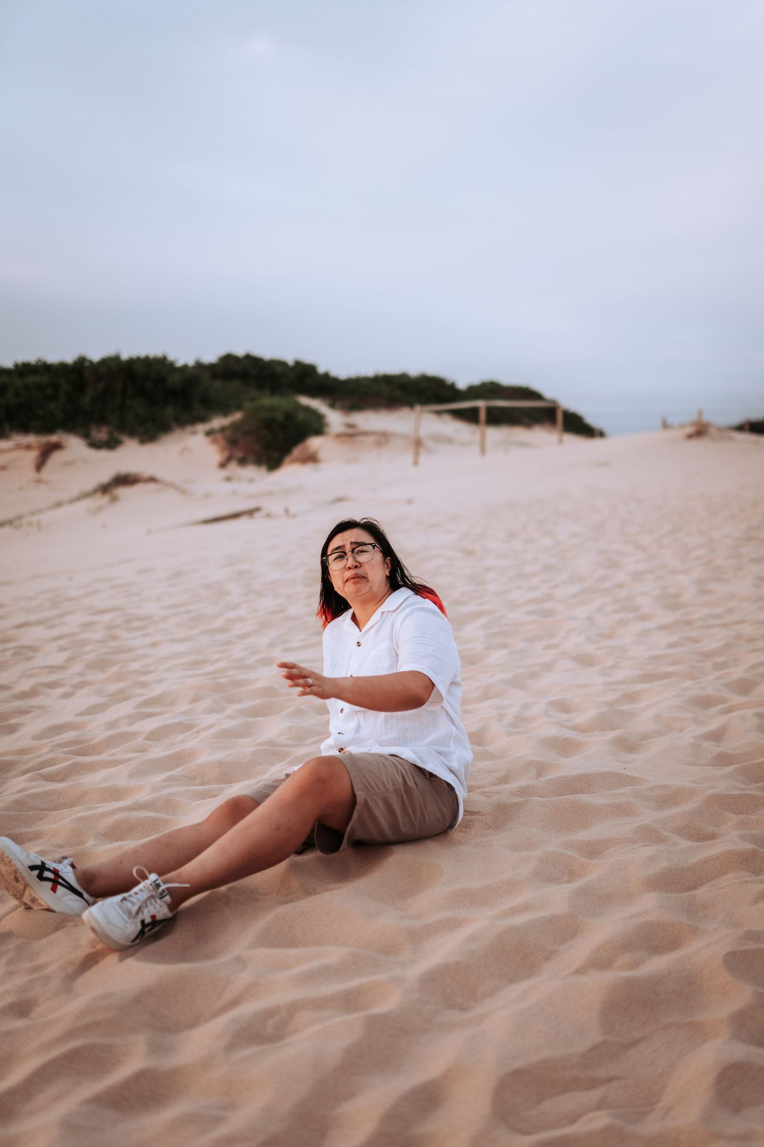 Person sitting on sandy dunes wearing white shirt and shorts against cloudy sky at sunset.