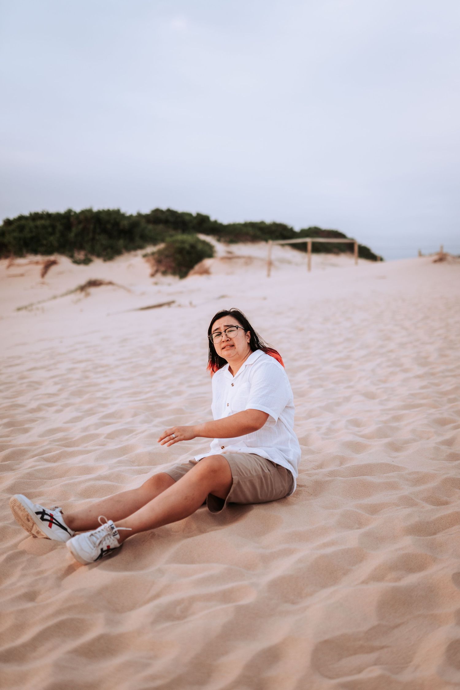 Someone wearing white shirt and shorts sits casually on sandy dunes at a beach during sunset.