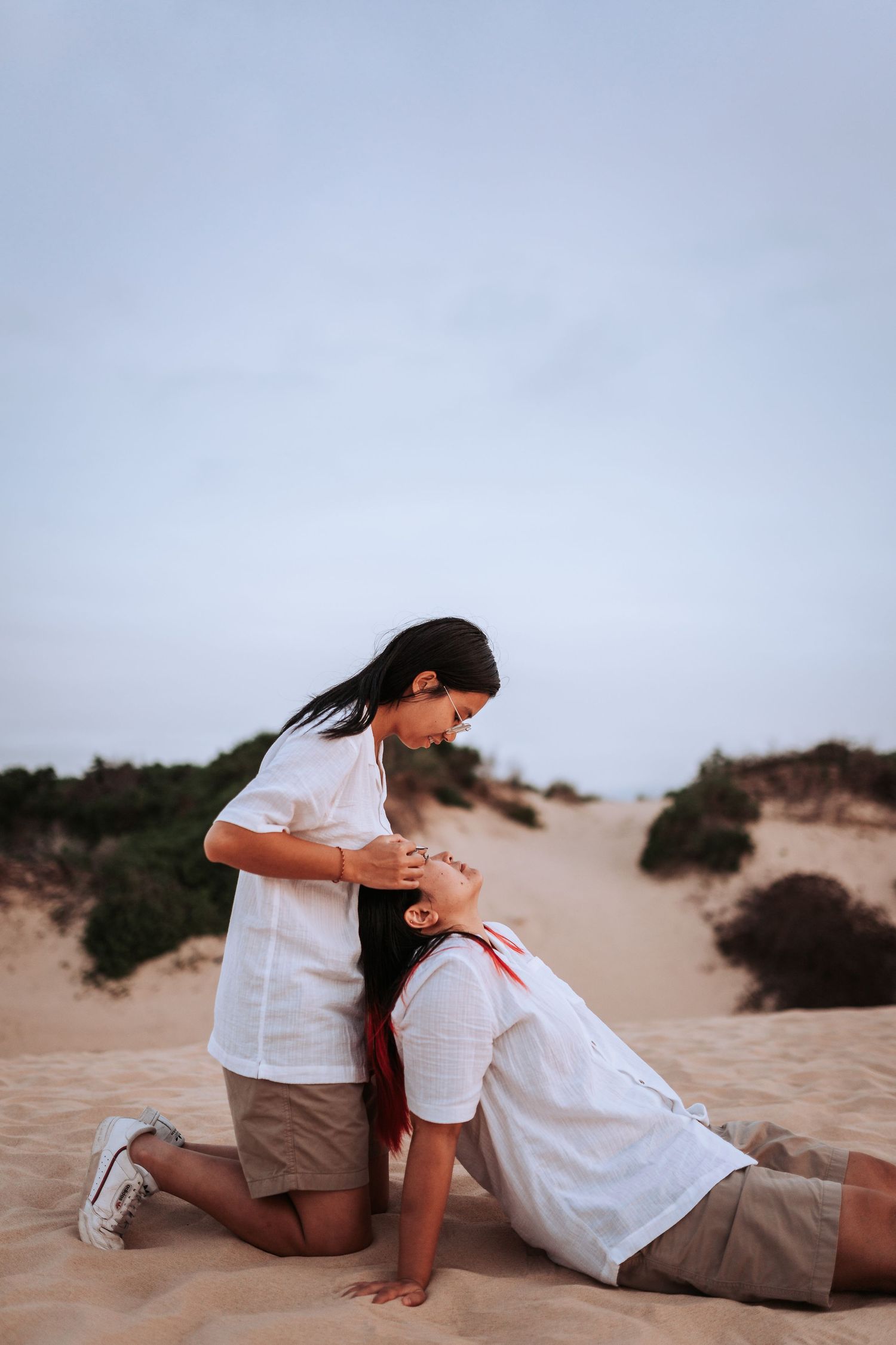Two people in white shirts share an intimate moment while kneeling on sand dunes at dusk.