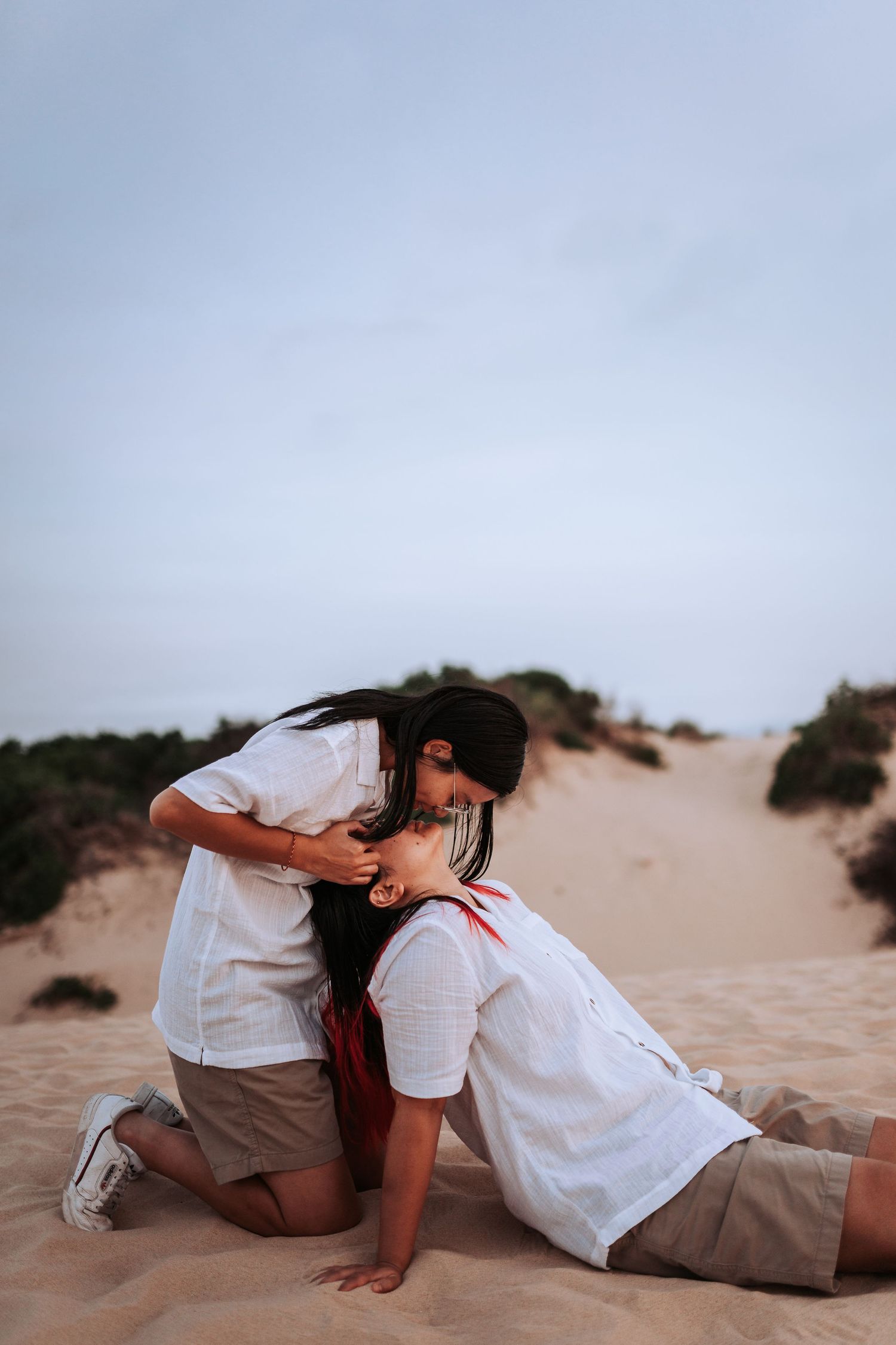 A romantic couple in white shirts share an intimate moment while sitting on sandy dunes at sunset.