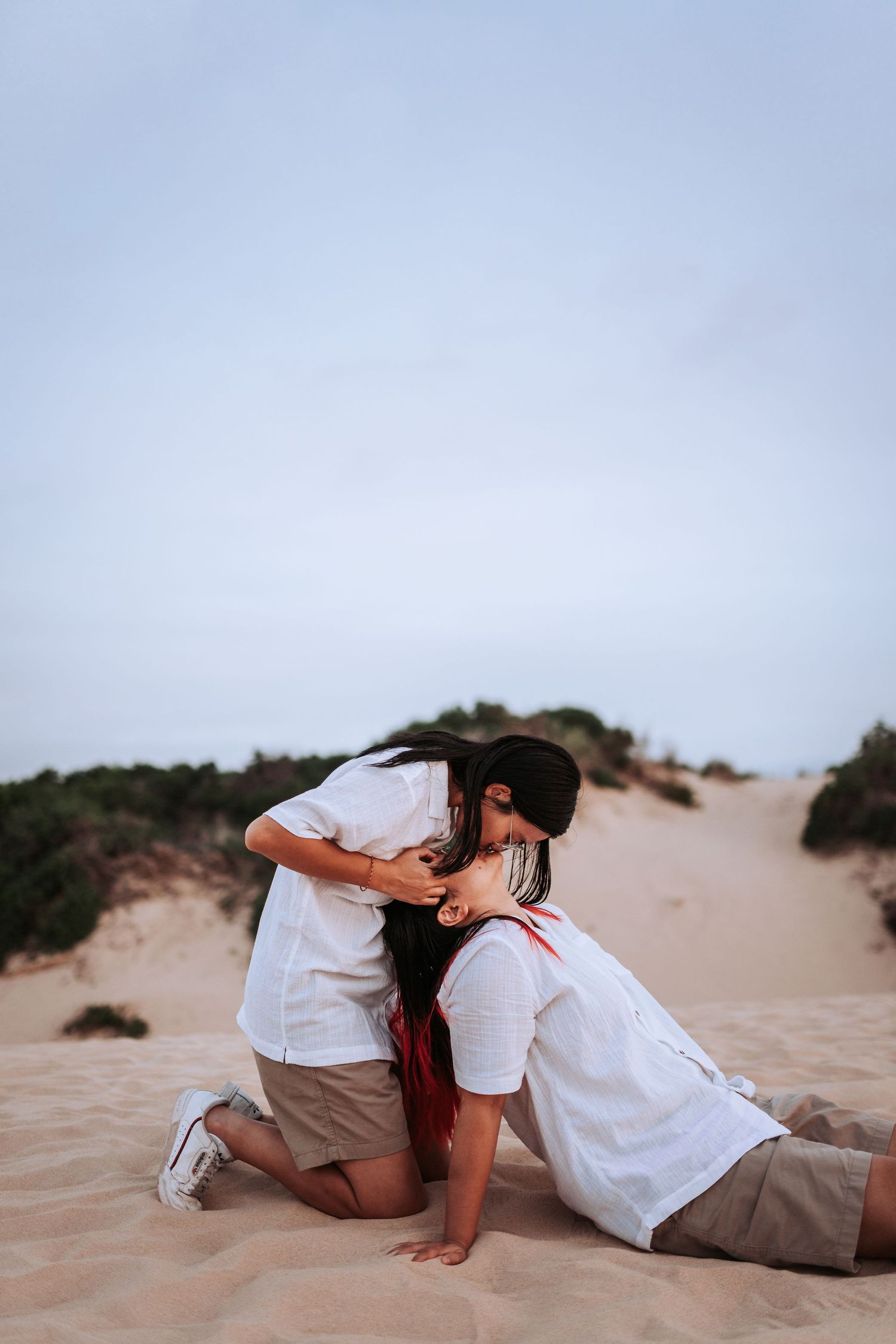 Two people in white shirts and khaki shorts share a playful moment while kneeling on sandy dunes at sunset.