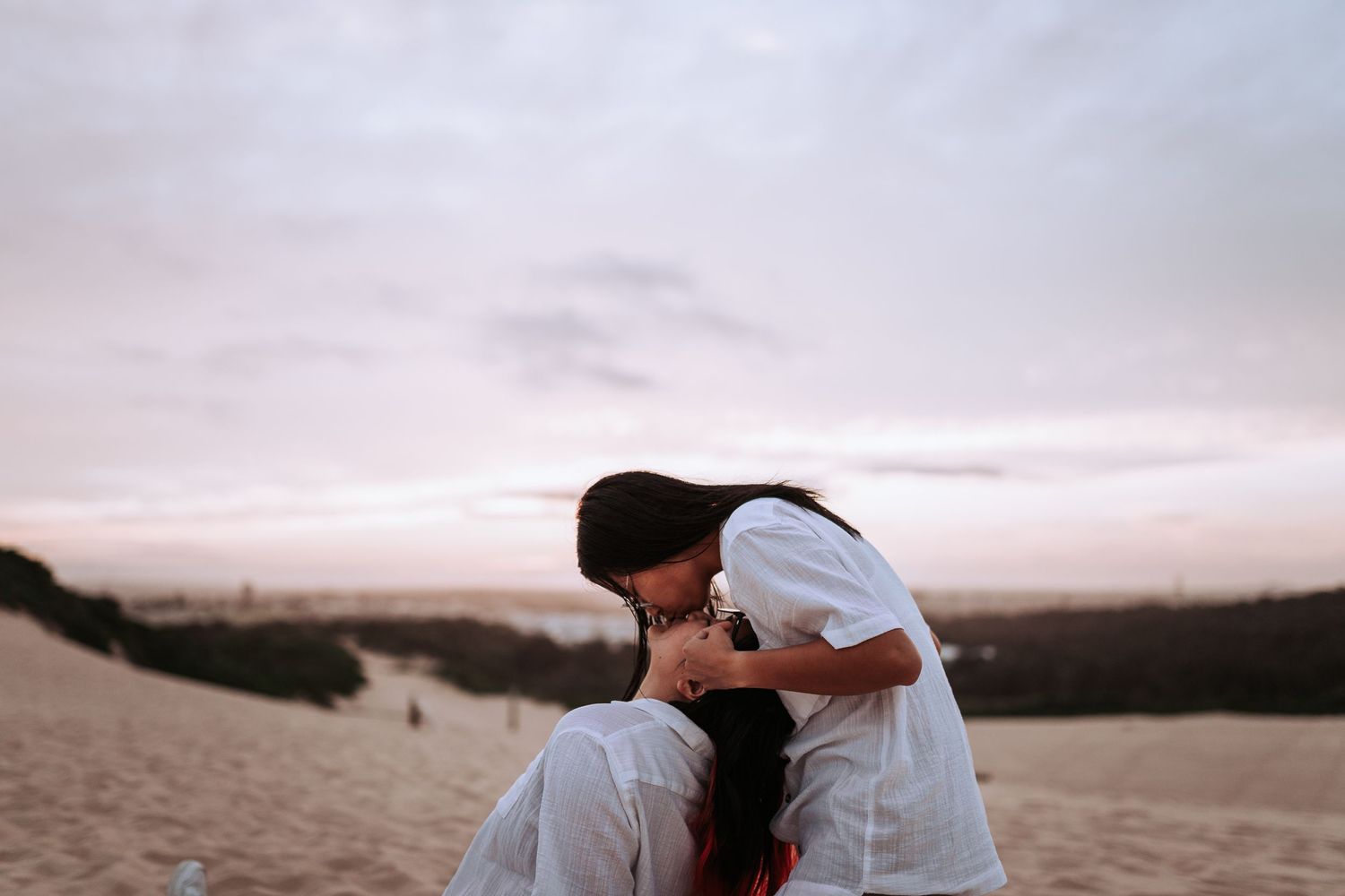 Two people share an intimate moment while sitting on sand dunes during a moody sunset.