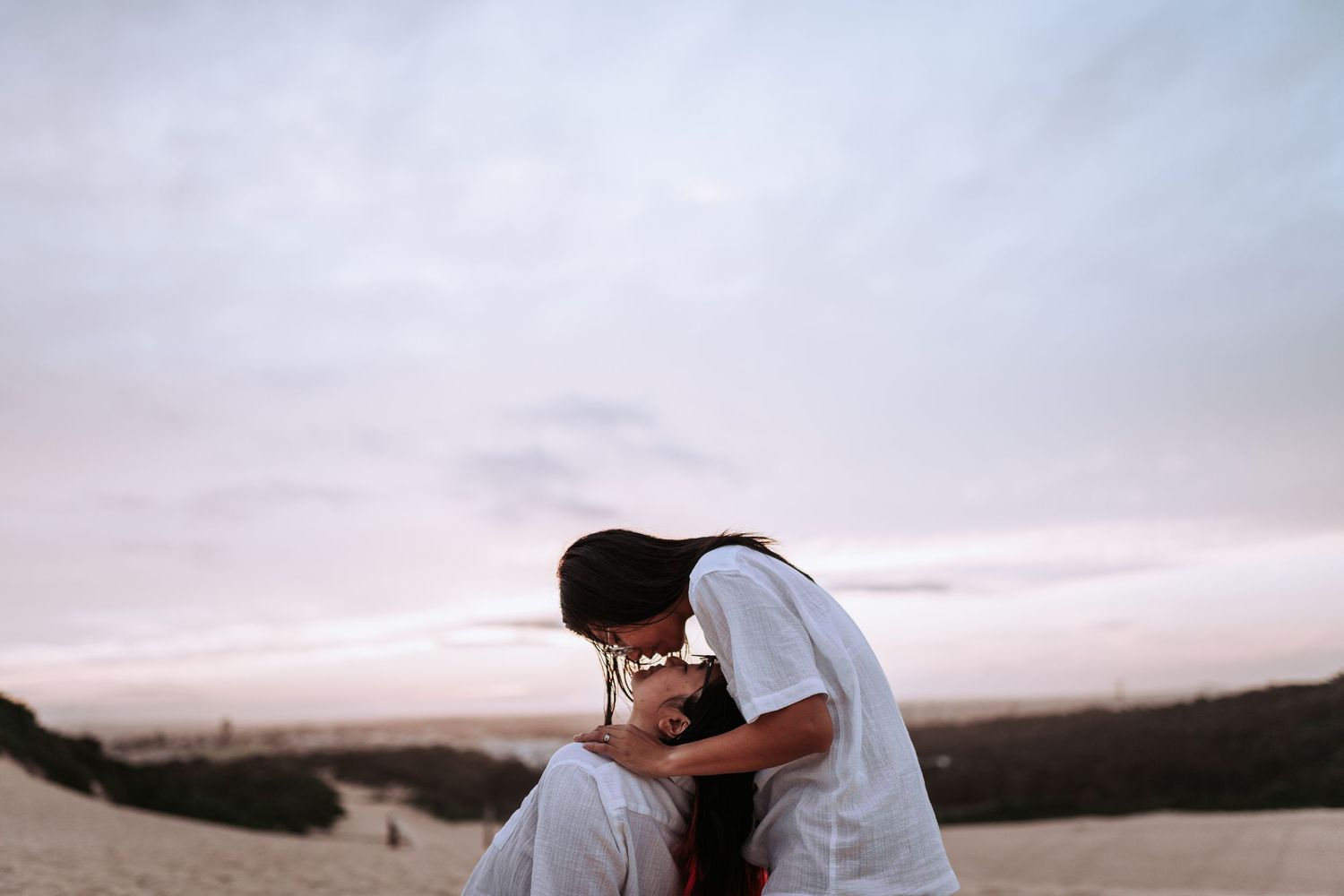 A romantic sunset moment between two people embracing on a desert dune with a soft pink sky in the background.