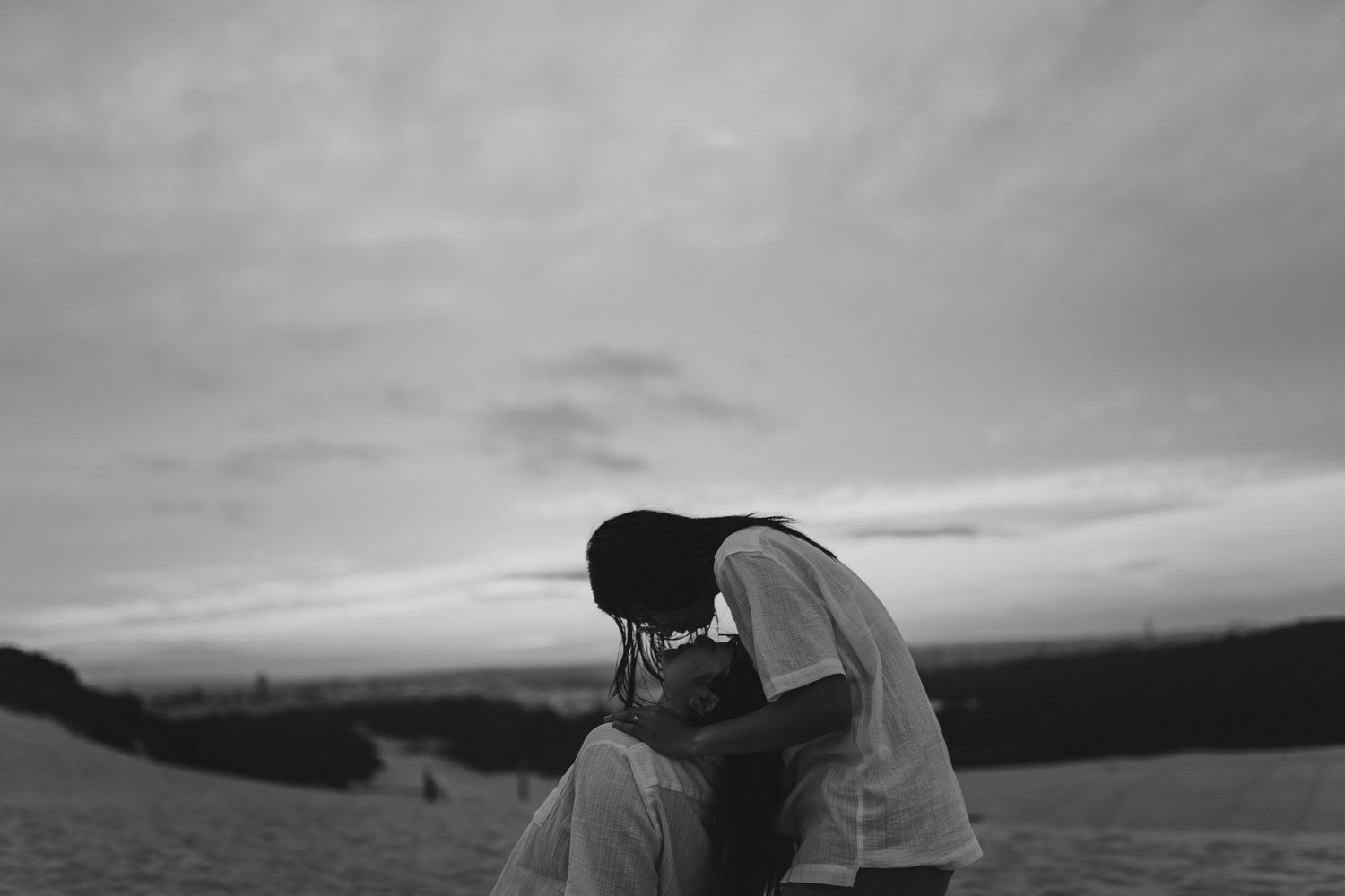 A couple silhouetted against cloudy skies share an intimate moment on a hilltop in black and white.