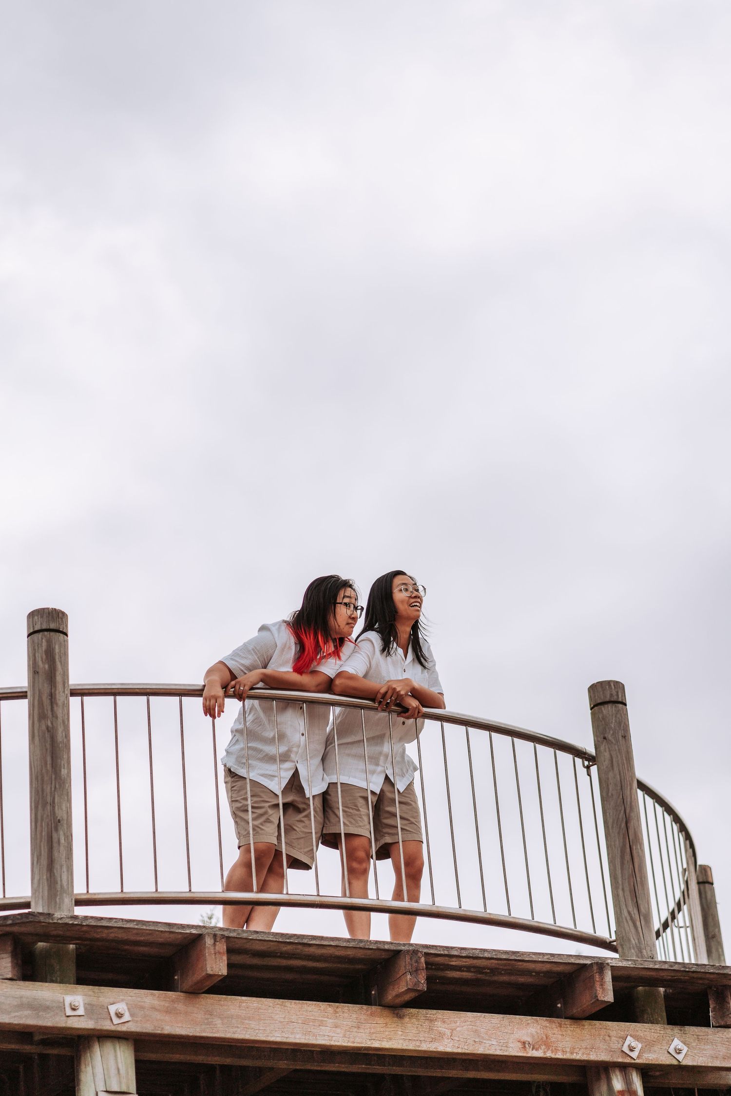 Two people in matching white shirts lean over a wooden railing on an elevated platform against a cloudy sky.