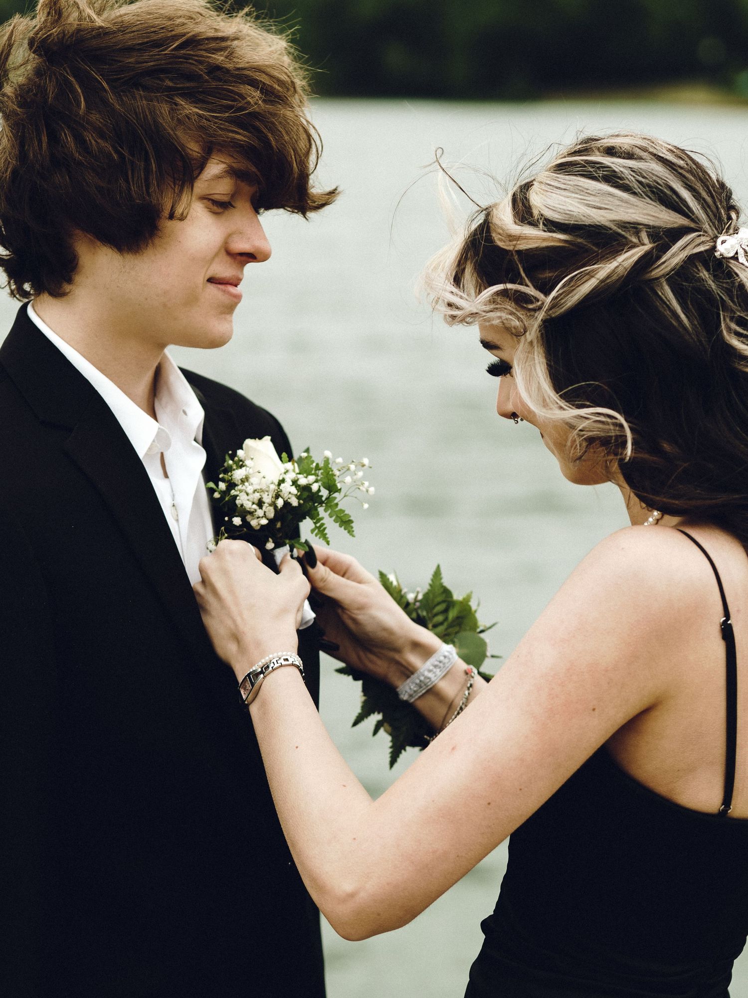 A couple at prom or formal event shares an intimate moment as she pins a white boutonniere flower on his black suit lapel.