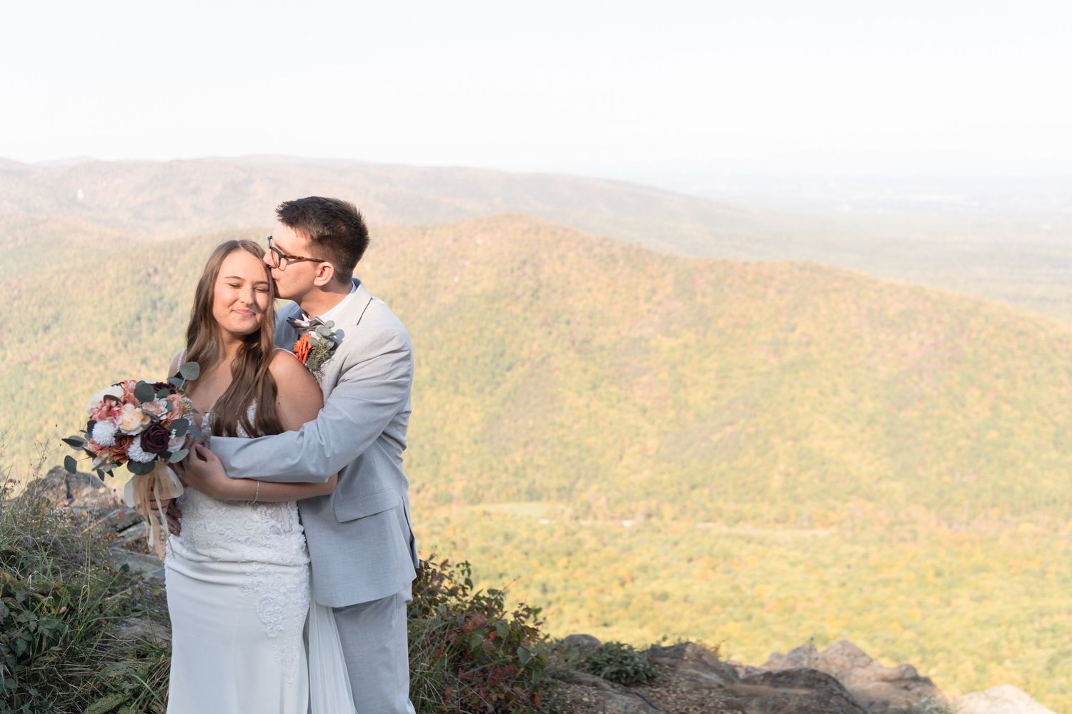 A couple in wedding attire embraces and laughs together on a scenic mountaintop overlook on their special day.