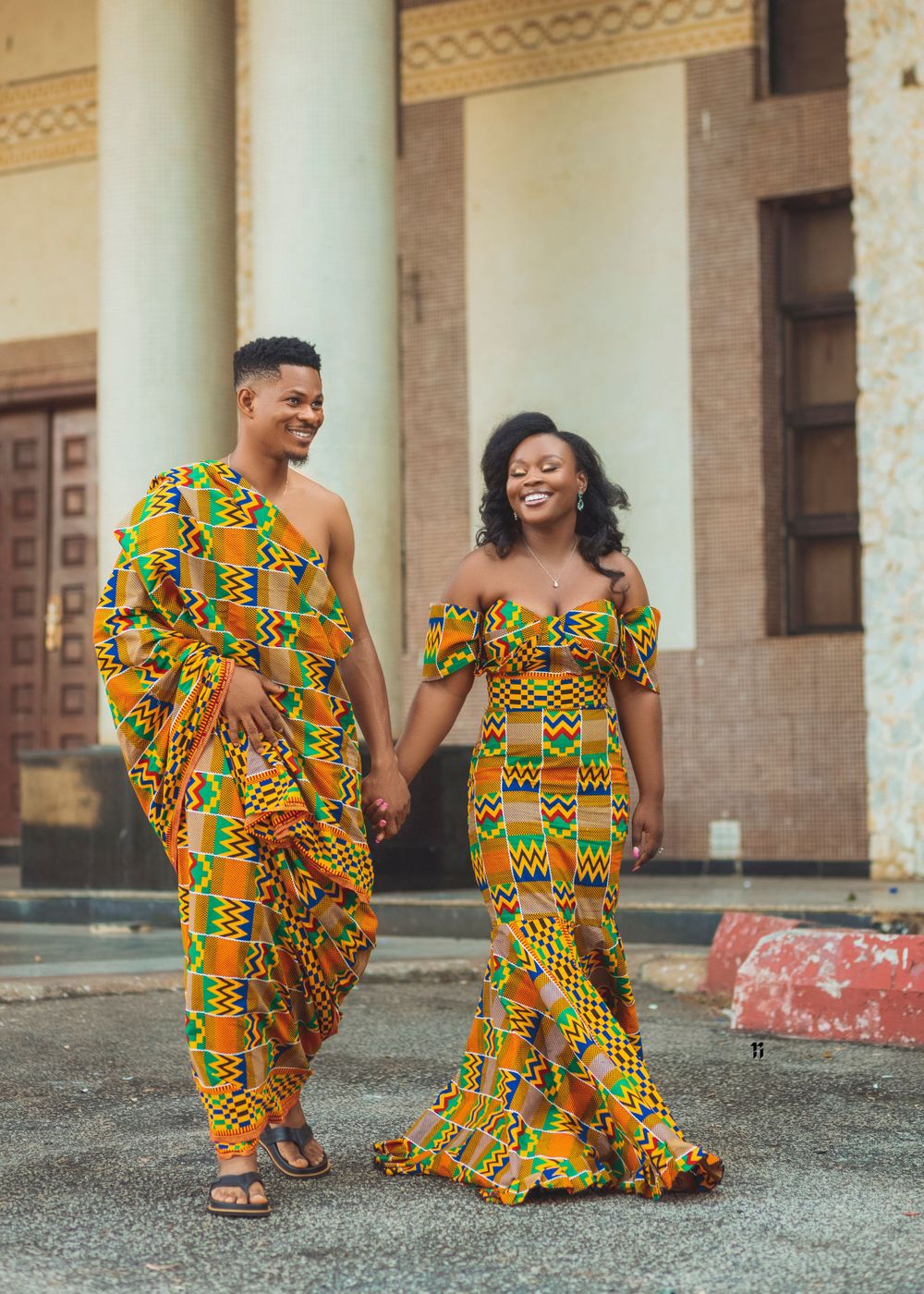 A couple wearing vibrant yellow and orange African Kente print outfits walk and laugh together outside a building.