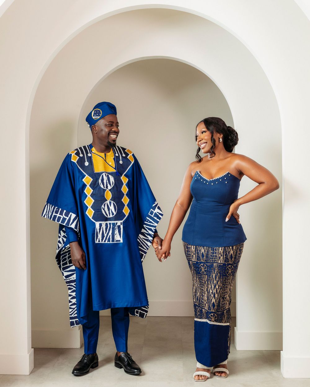 A couple in traditional African attire poses in an archway, wearing coordinating blue and yellow formal ethnic clothing.
